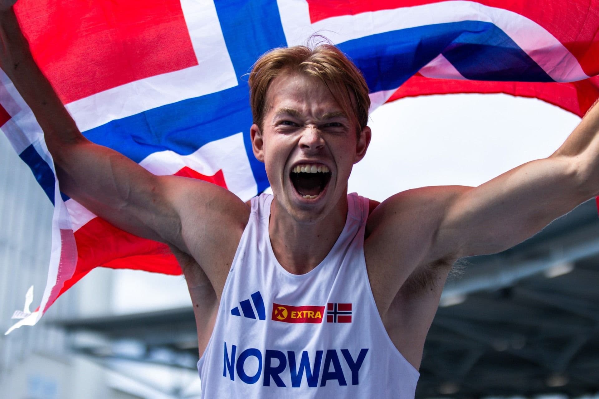 En jublende friidrettsutøver i hvit landslagssinglet med teksten «Norway» løfter et norsk flagg bak seg.
