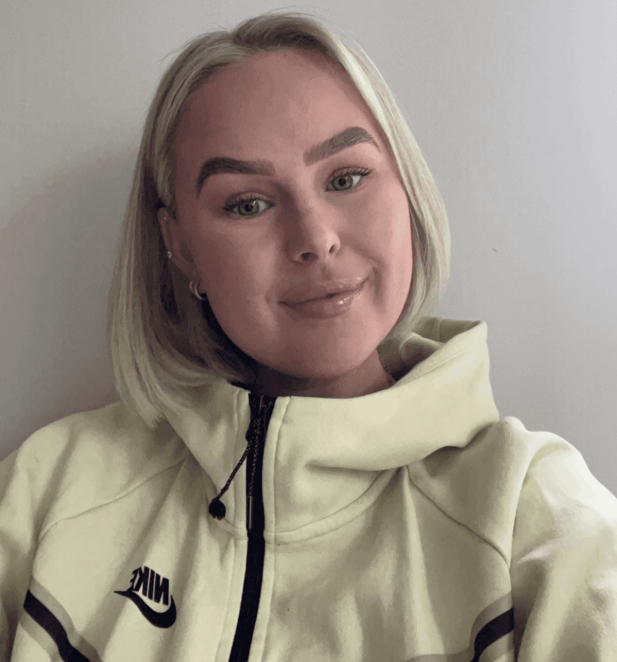 Portrett av en blond kvinne i en lys Nike-hettejakke mot en lys vegg.