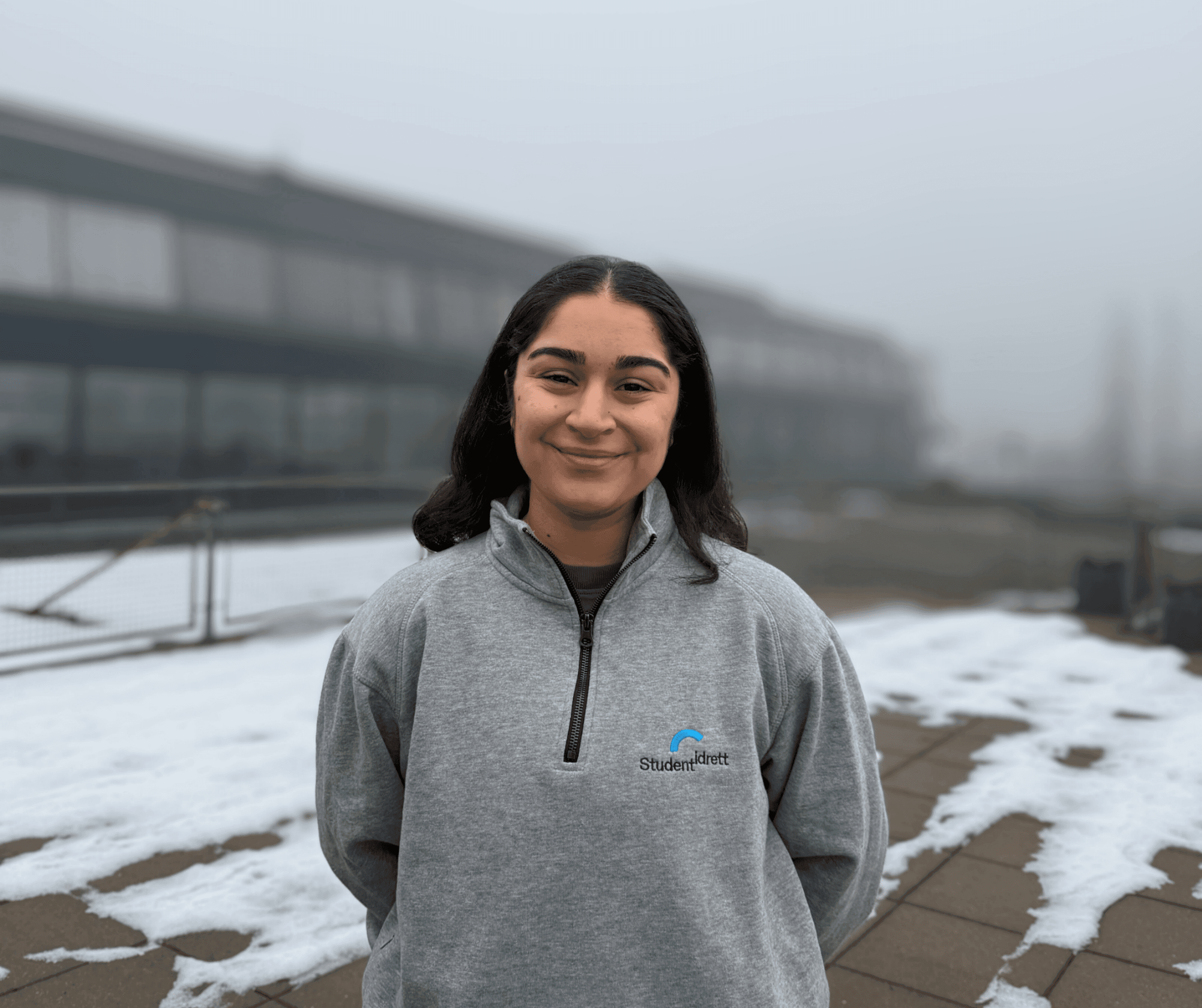 En smilende ung kvinne i grå Studentidrett-genser står utenfor en campusbygning med snøflekker på bakken.