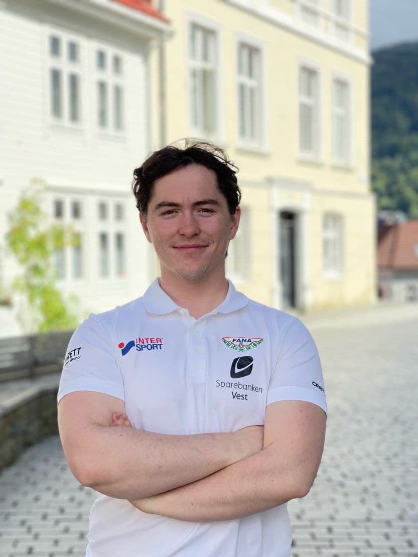 En ung mann med korslagte armer i hvit poloskjorte med klubblogo, foran en lys bygning.