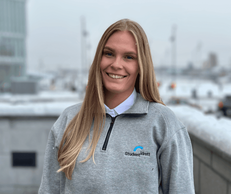 Portrett av en smilende kvinne i grå Studentidrett-genser med snødekte tak i bakgrunnen.