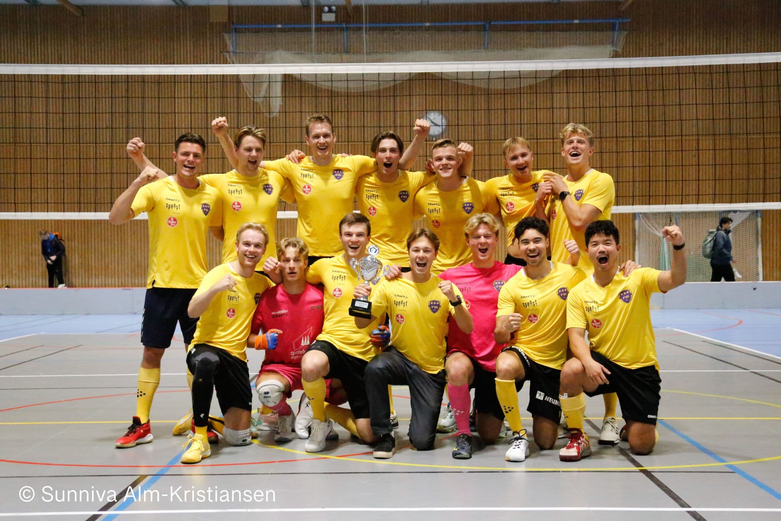 Et futsallag i gule drakter jubler med en pokal i en idrettshall.