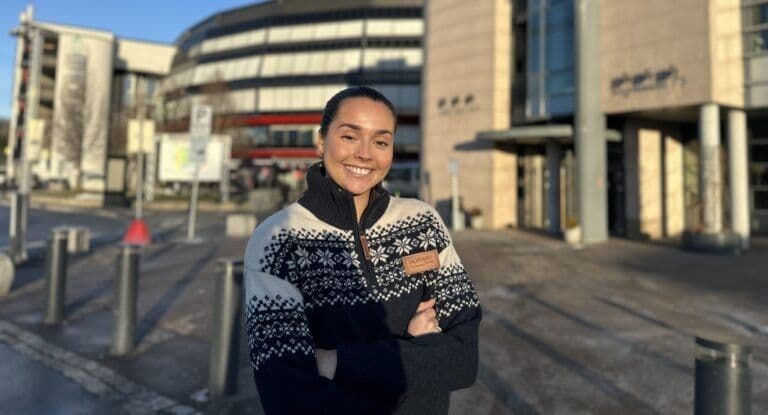 Smilende kvinne i norsk mønstergenser står på en byplass foran et kjøpesenter i sollys.