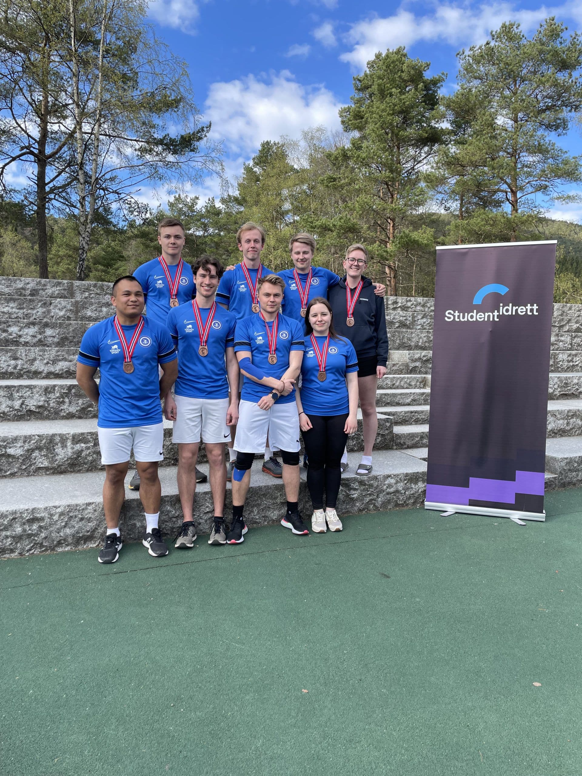 En gruppe studenter i blå drakter med medaljer poserer foran en Studentidrett-rollup utendørs.