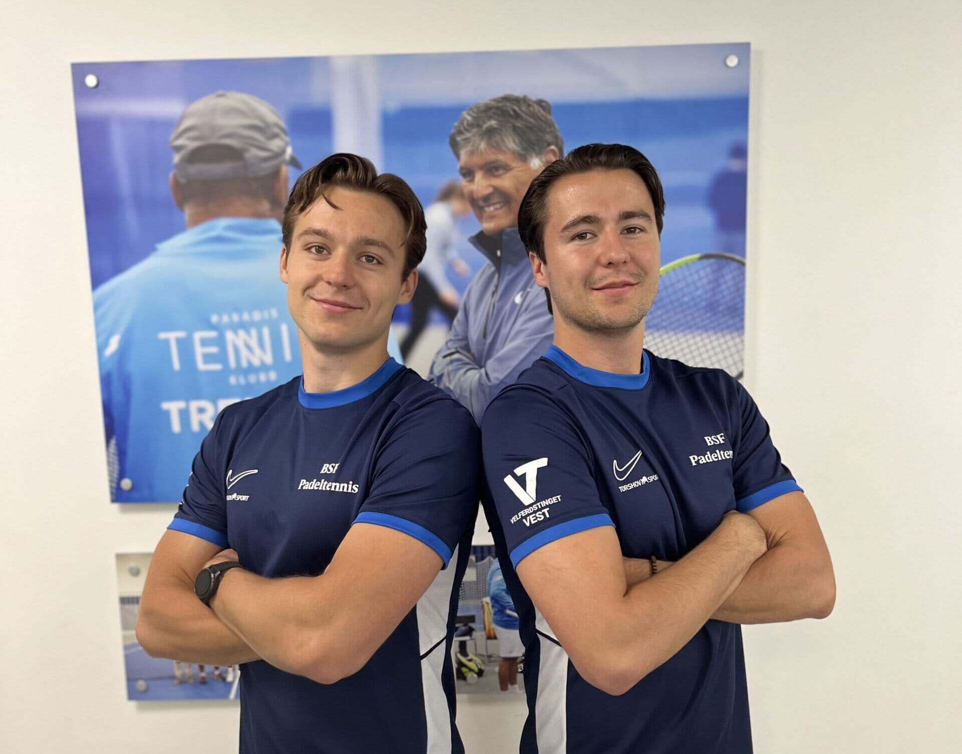 To smilende menn i blå padeltennis-drakter står med korslagte armer foran en tennisplakat.