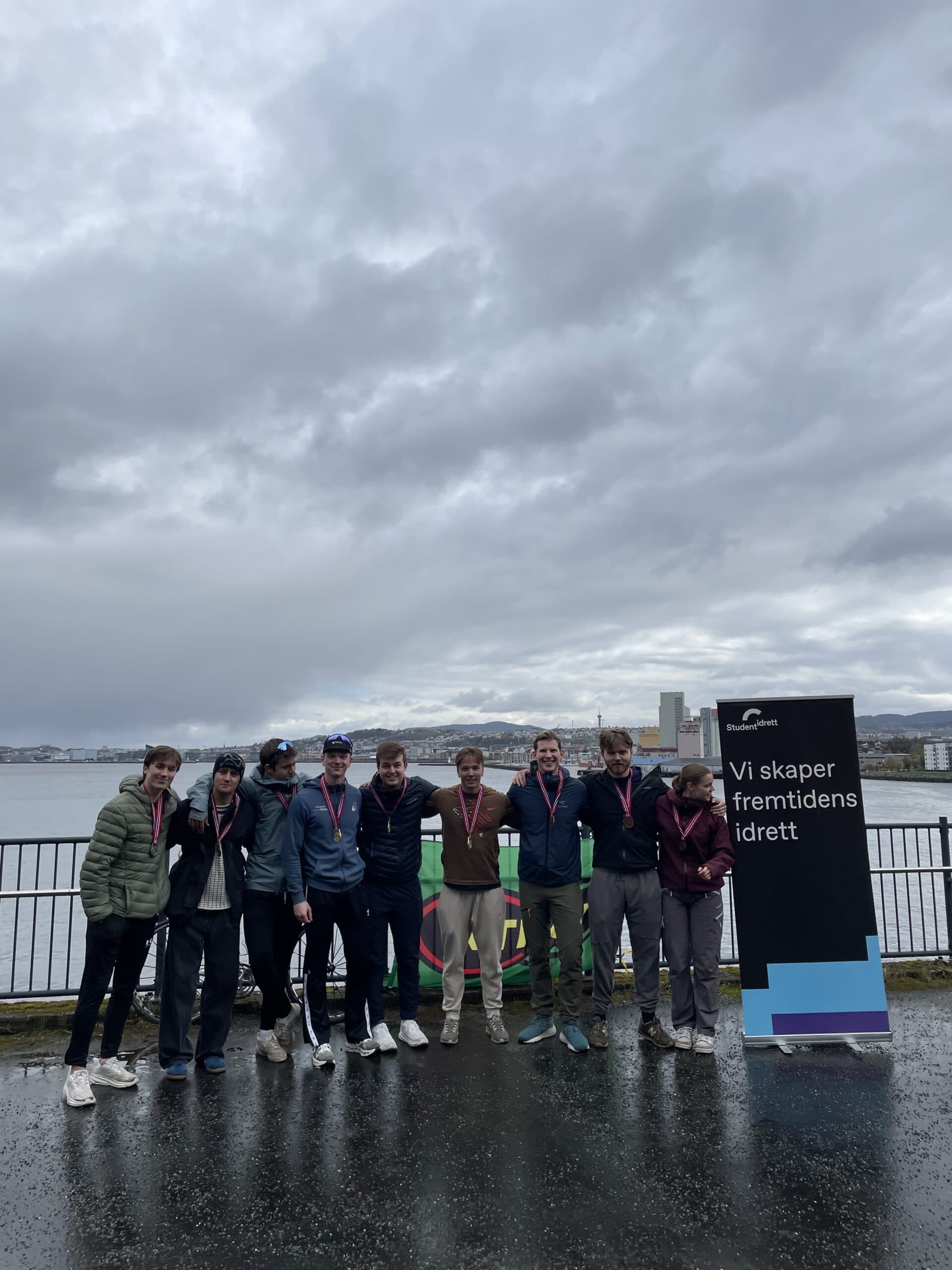 En gruppe unge menn og en kvinne med medaljer poserer ved sjøen foran en Studentidrett-rollup med teksten Vi skaper fremtidens idrett.