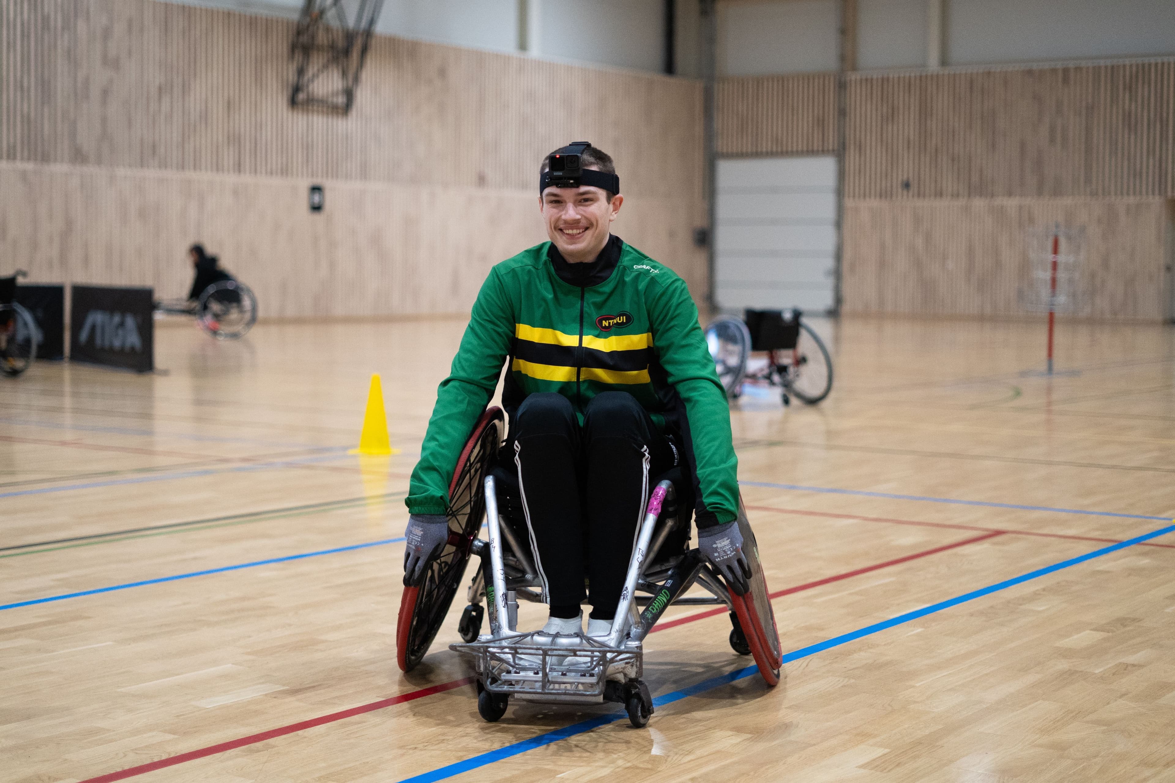 En smilende utøver i kjelkeidrett med grønn drakt kjører over et idrettsgulv i en hall.