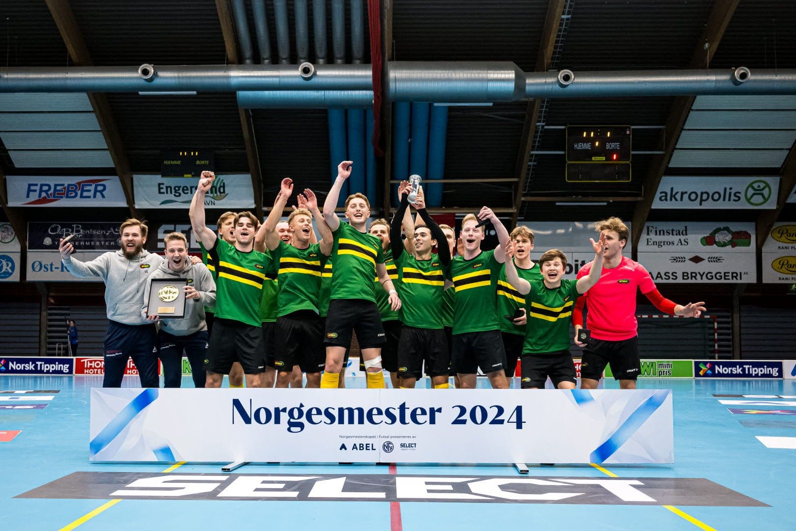Et jublende herrelag i grønne og gule drakter feirer seier foran en skjerm med teksten Norgesmester 2024.