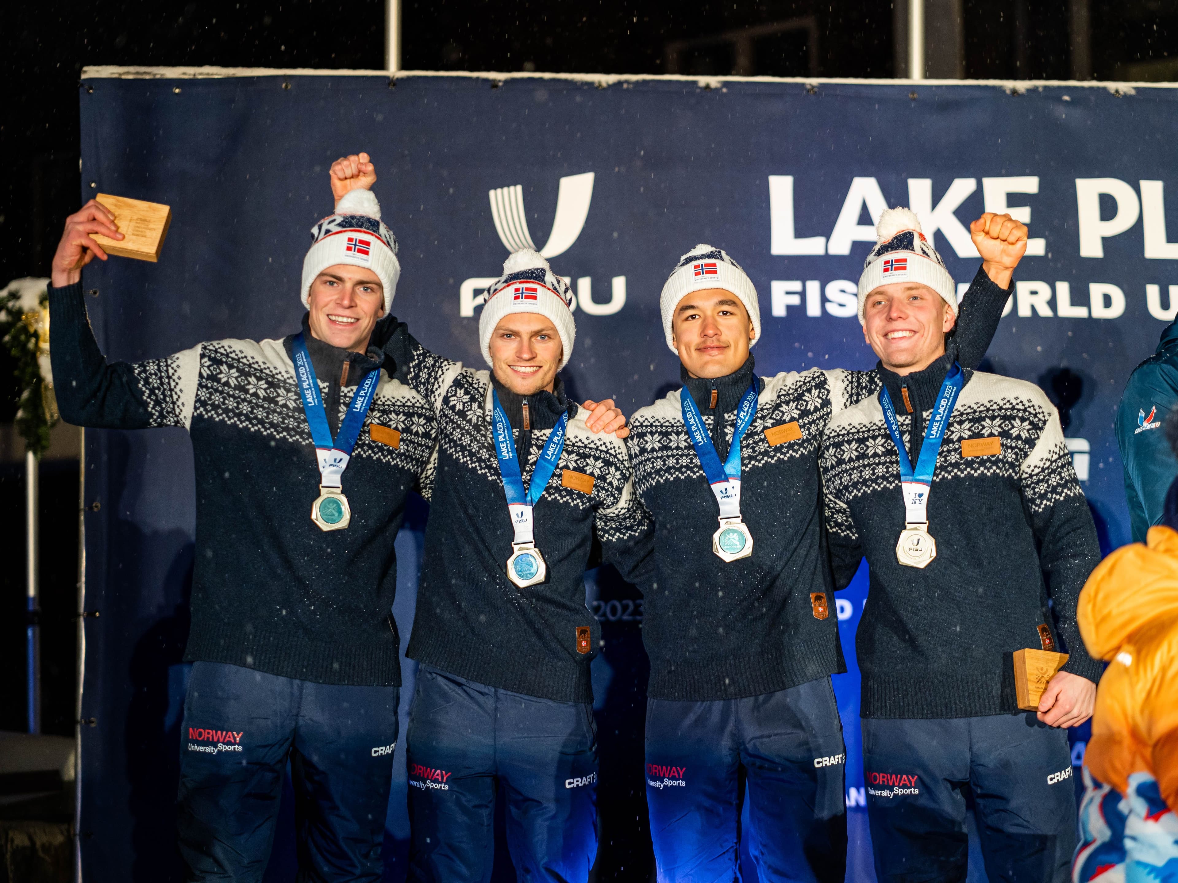 Fire mannlige langrennsløpere med medaljer og norske luer jubler på pallen under FISU World University Games i Lake Placid.