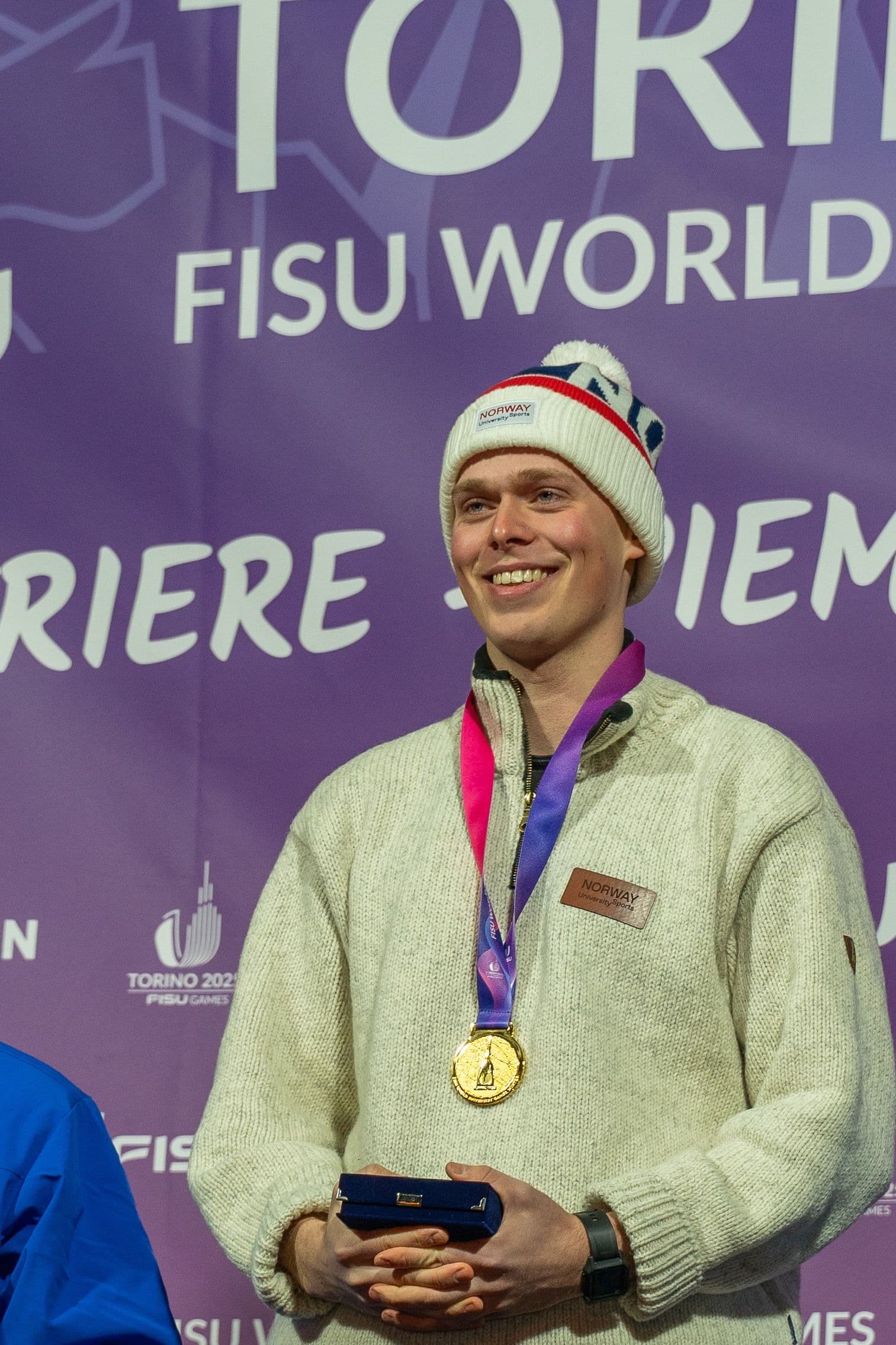 En smilende mann med lue og gullmedalje foran en vegg merket FISU World University Games Torino 2025.