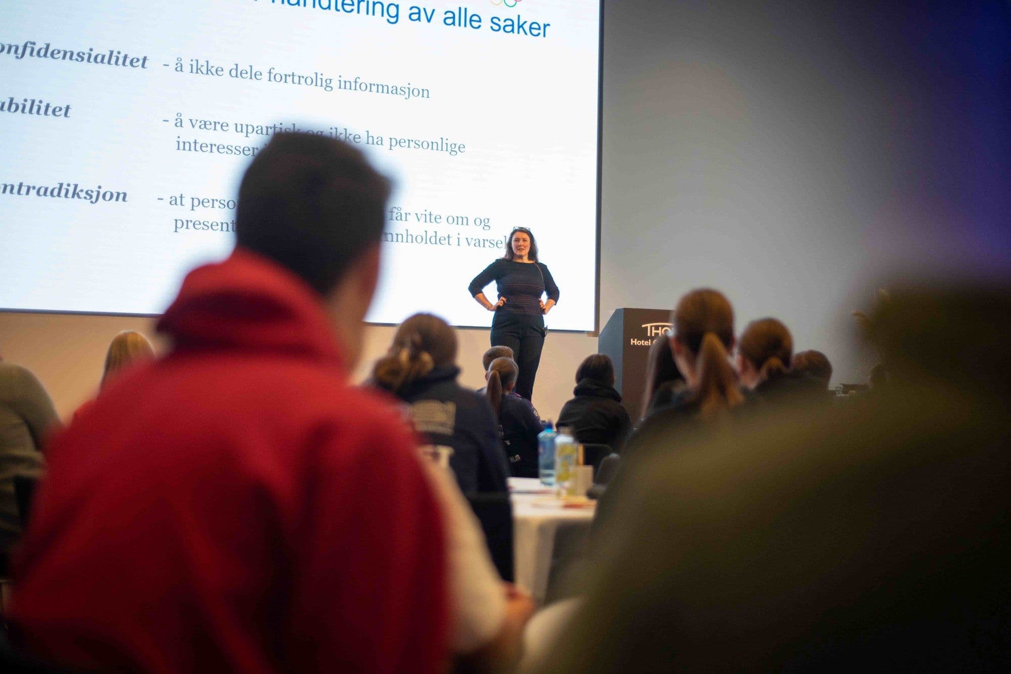 En kvinne holder foredrag foran et publikum med en presentasjon om konfidensialitet og håndtering av saker på lerretet.