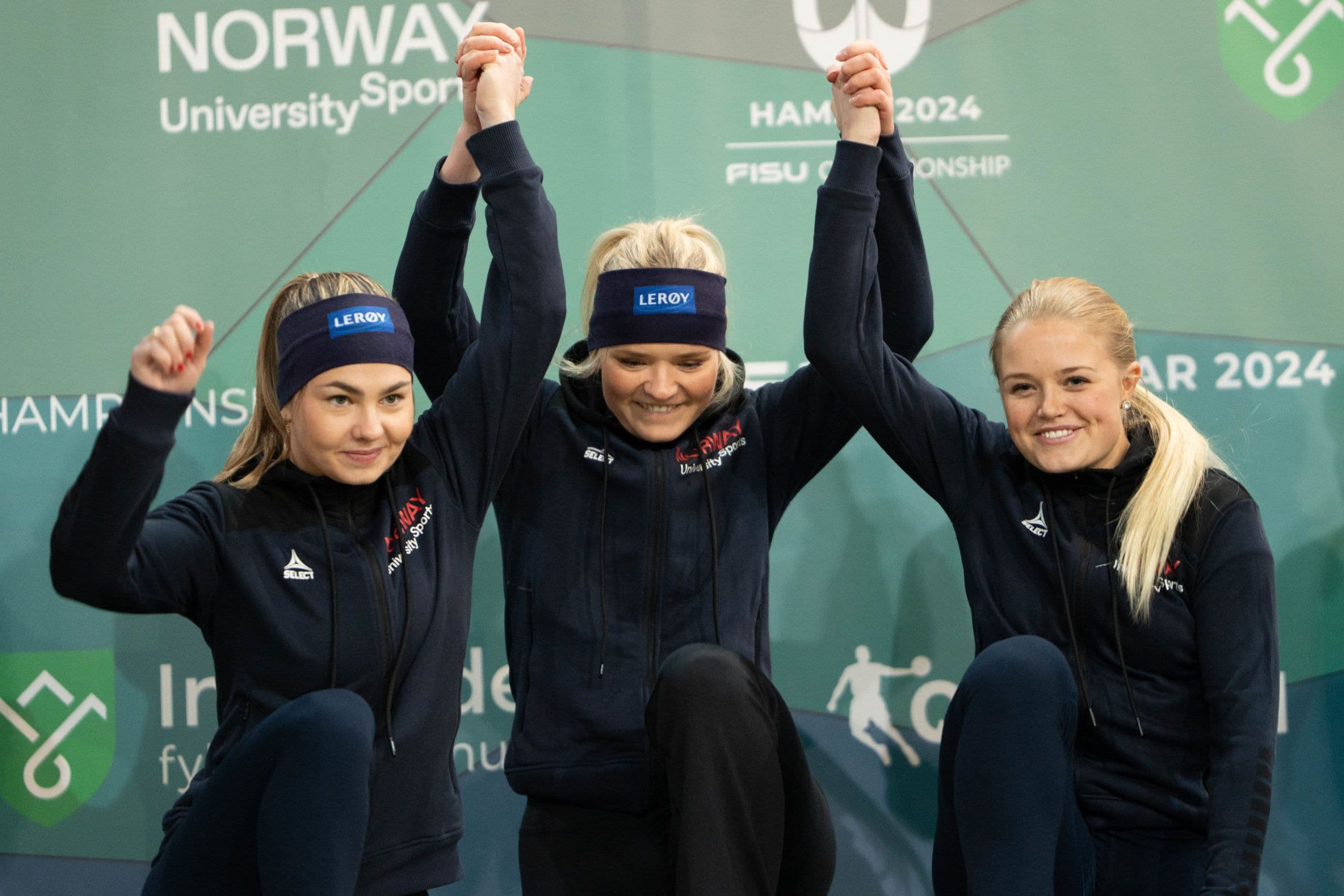 Tre kvinnelige utøvere i Norway-drakter løfter hendene sammen foran en vegg merket Hamar 2024 FISU Championship.