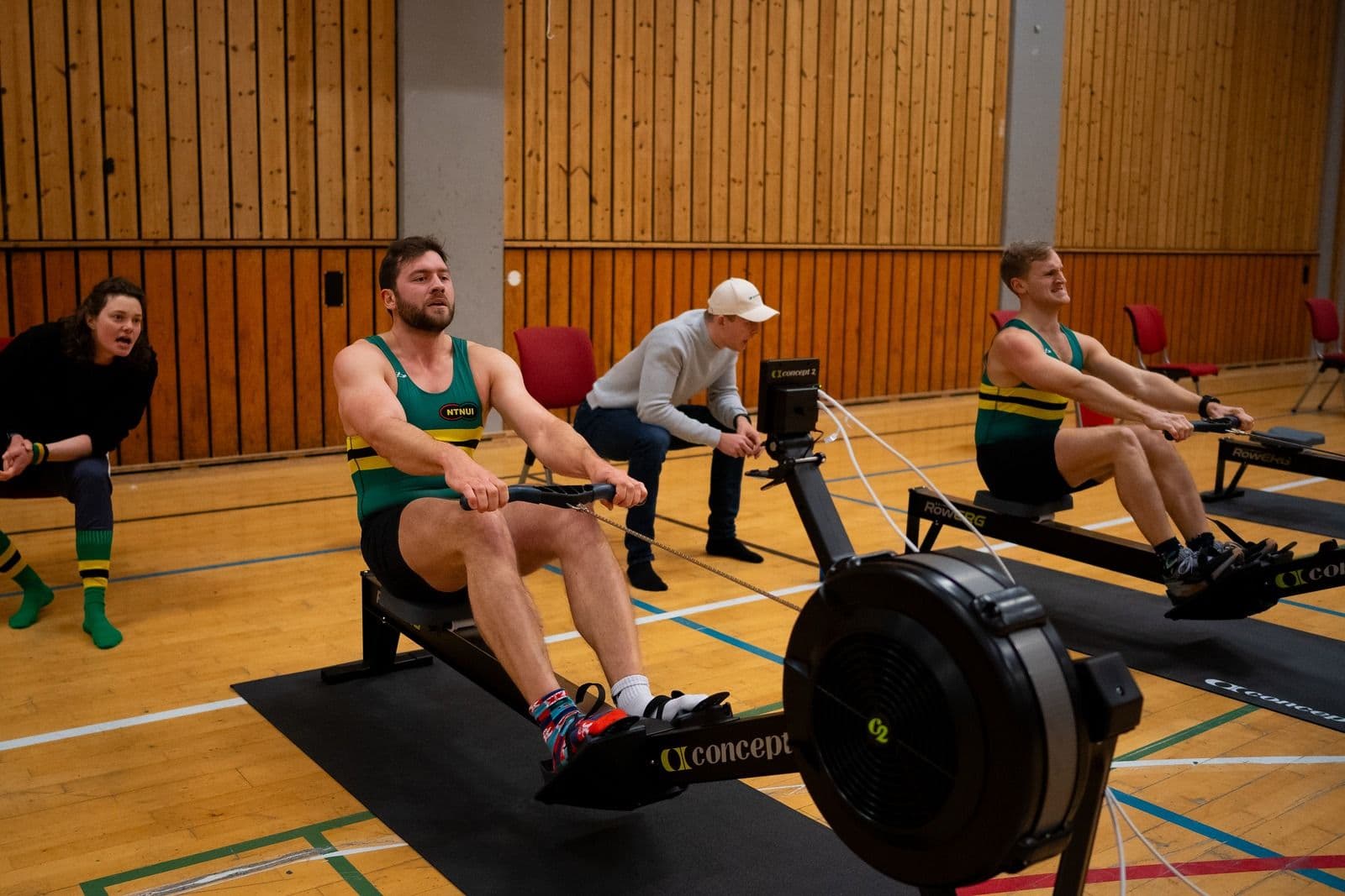 Flere personer ror på romaskiner i en gymsal mens en kvinne heier på siden.