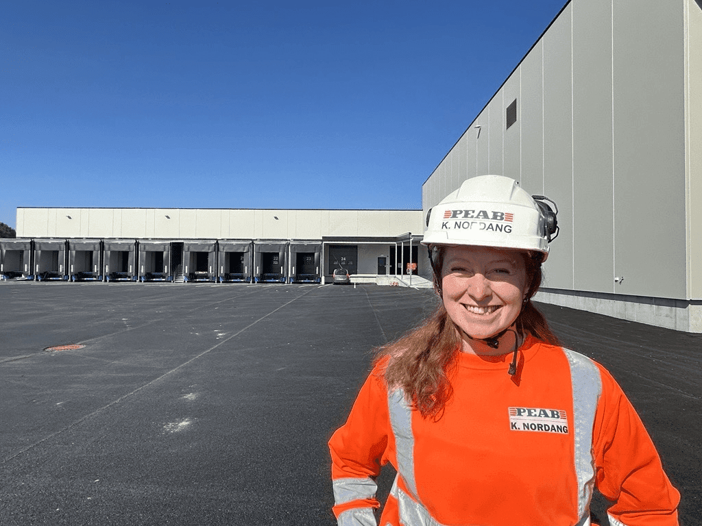 En kvinne i oransje arbeidsklær og hjelm smiler foran et stort næringsbygg under en klar blå himmel.