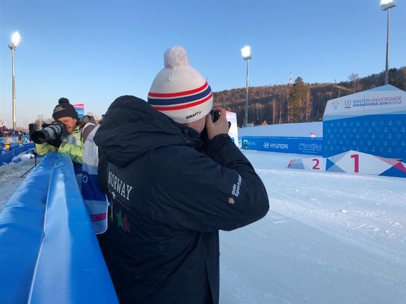 En mann i Norway-jakke og lue fotograferer ved en langrennsarena under Winter Universiade Krasnoyarsk 2019.