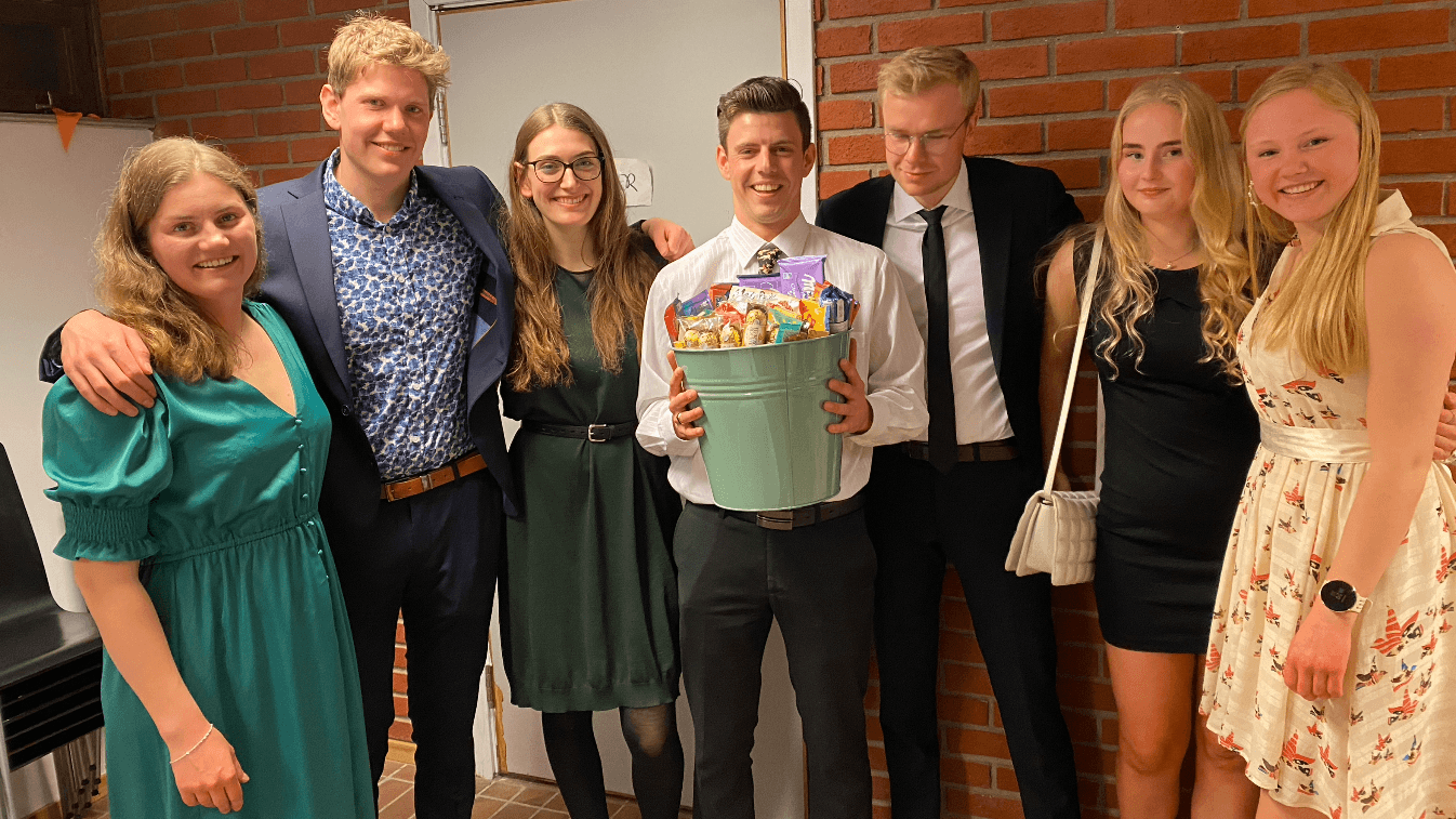 En gruppe unge personer i penklær smiler sammen, der en av dem holder en bøtte fylt med godteri.