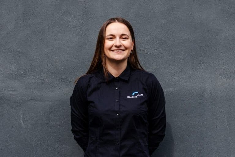 Portrett av en smilende kvinne i sort Studentidrett-skjorte mot en grå vegg.