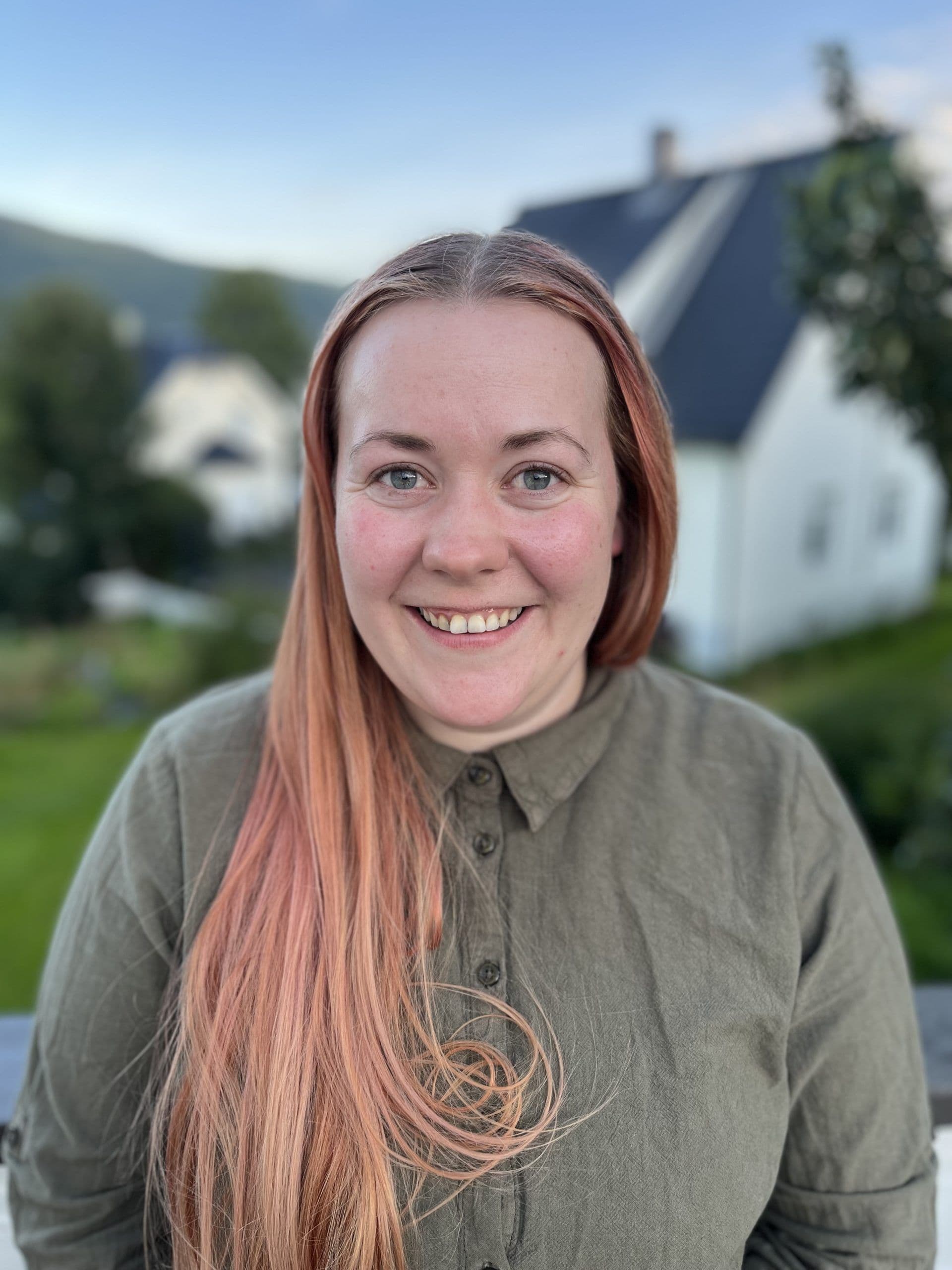 Portrett av en smilende kvinne med langt rødt hår i grønn skjorte, fotografert utendørs.