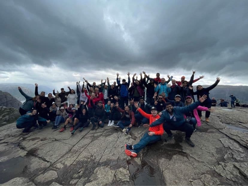 En stor gruppe studenter poserer med armene i været på en fjellplatå under en overskyet himmel.