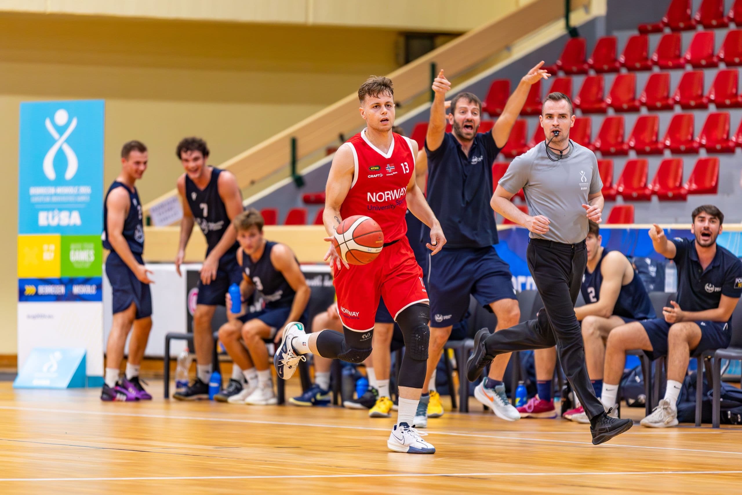 En basketspiller i rød Norway-drakt fører ballen under en kamp mens motspillere og dommer følger med.