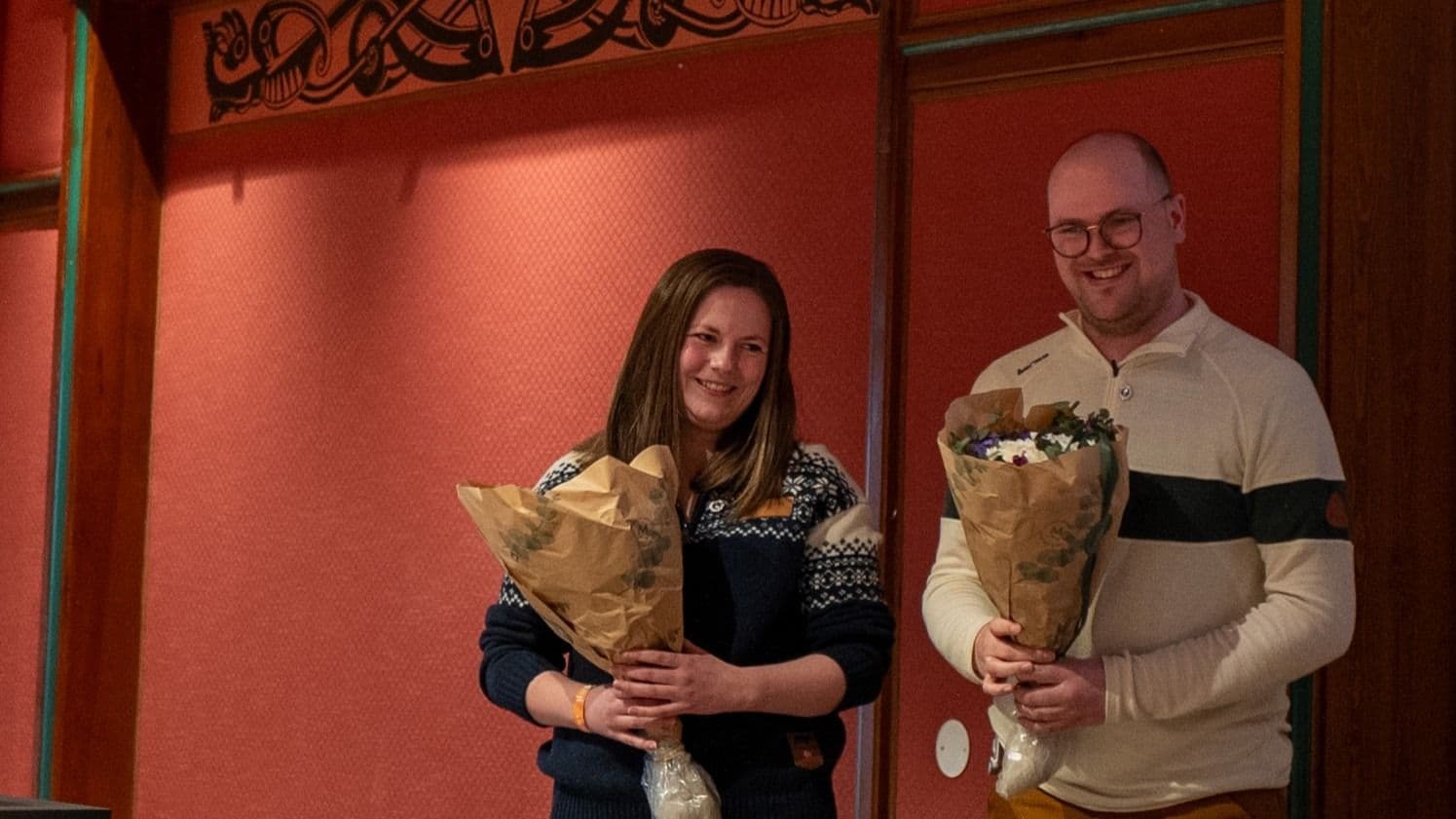 En kvinne og en mann står ved siden av hverandre og holder hver sin blomsterbukett foran en rødmalt vegg.