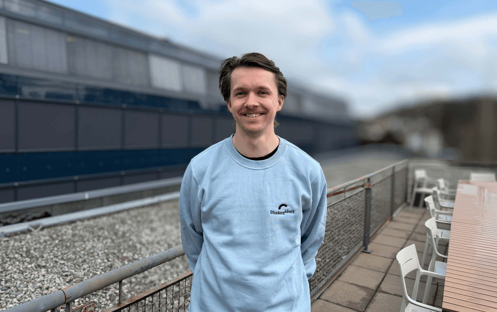 Portrett av en smilende mann i lyseblå Studentidrett-genser på en takterrasse.