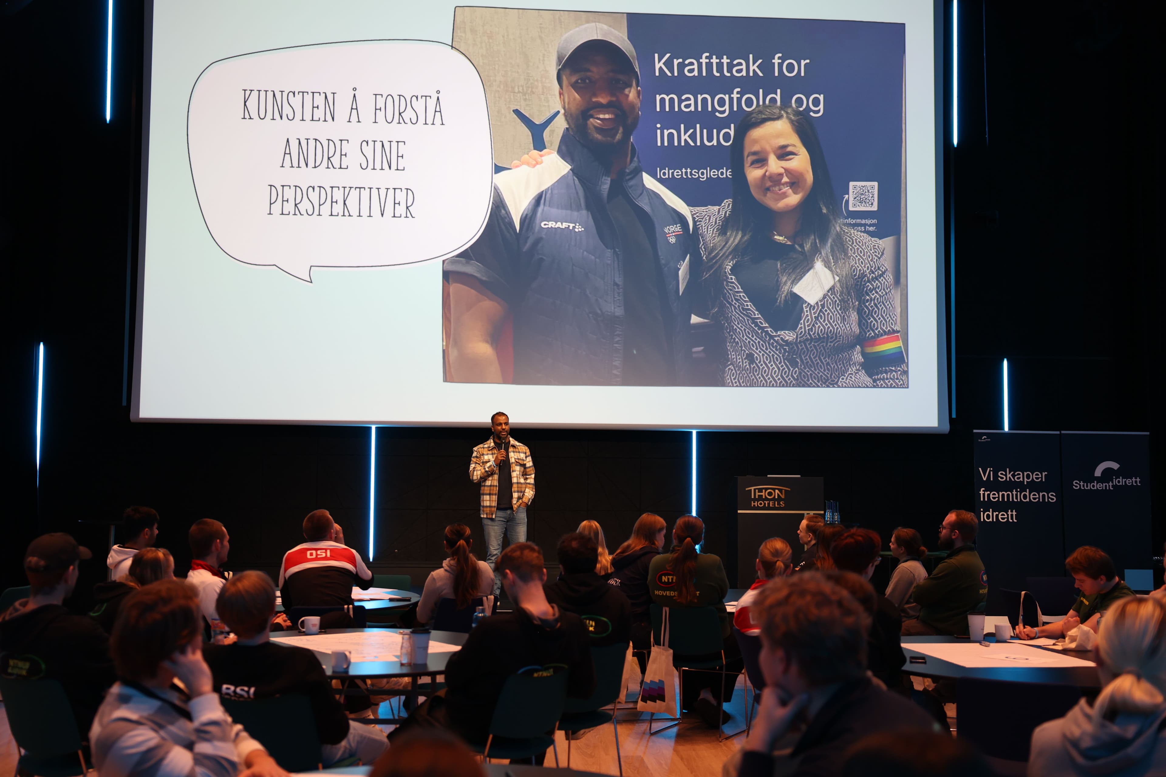 En person står på scenen foran en presentasjon med teksten «Kunsten å forstå andre sine perspektiver» foran et publikum.