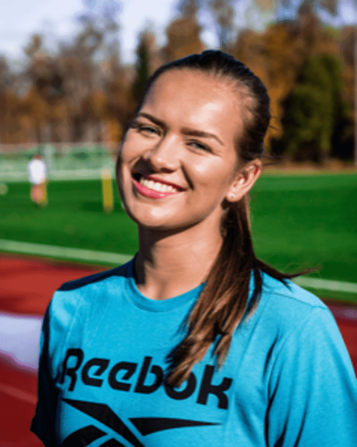 Portrett av en smilende kvinne i turkis Reebok-genser på et idrettsanlegg.