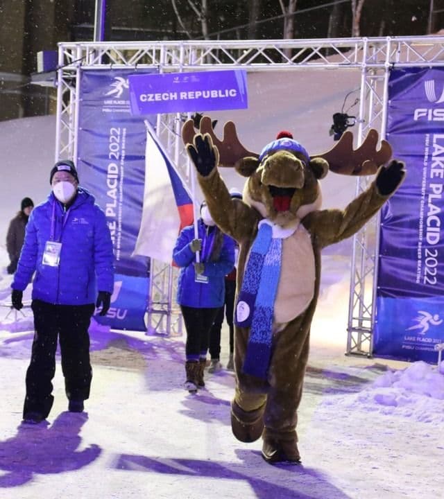 En elgmaskott med skjerf løfter armene under en nasjonsparade i snøen ved FISU World University Games i Lake Placid 2022, med en frivillig ved siden.