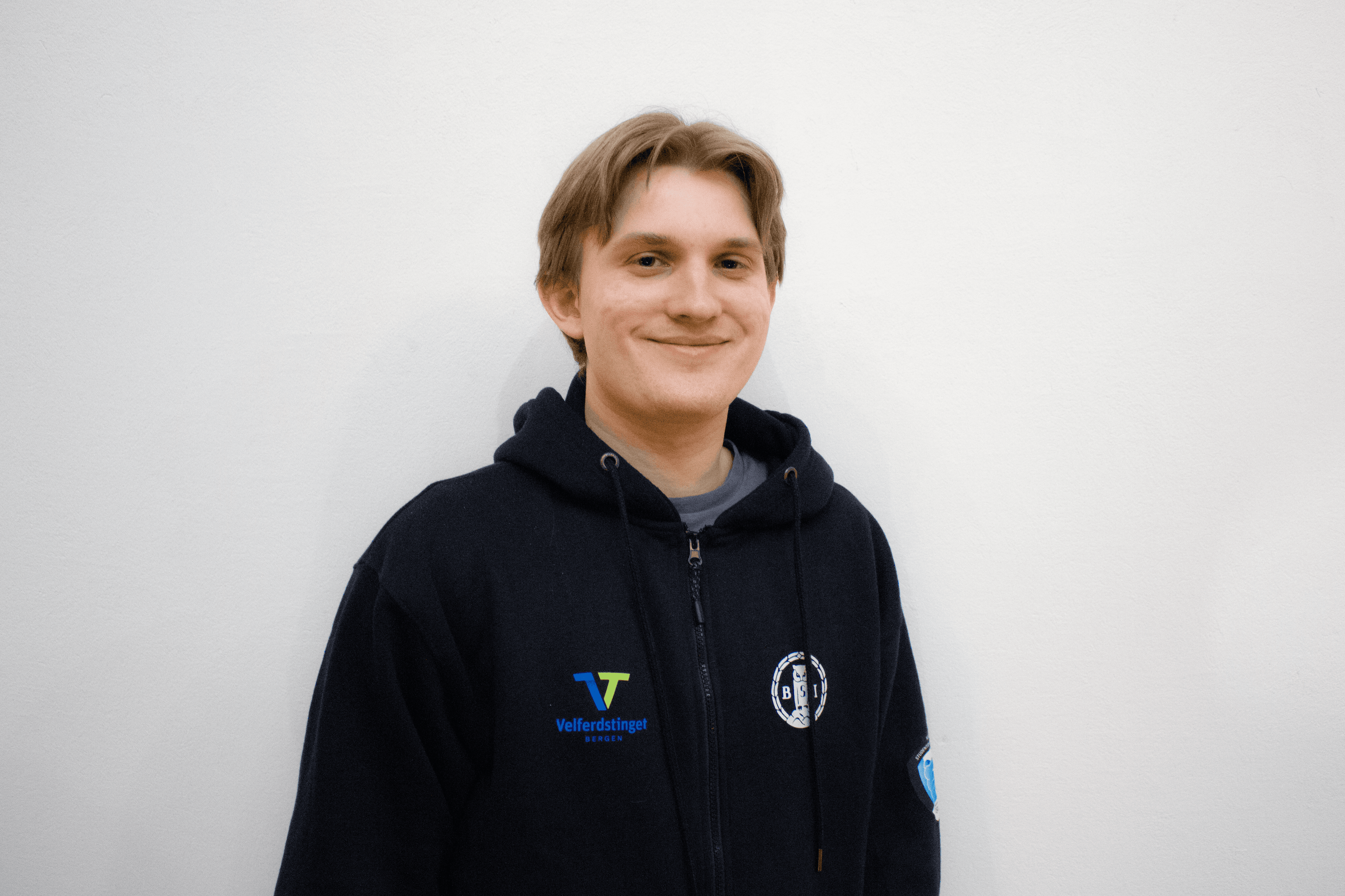 Portrett av en smilende mann i svart hettejakke mot en lys vegg.
