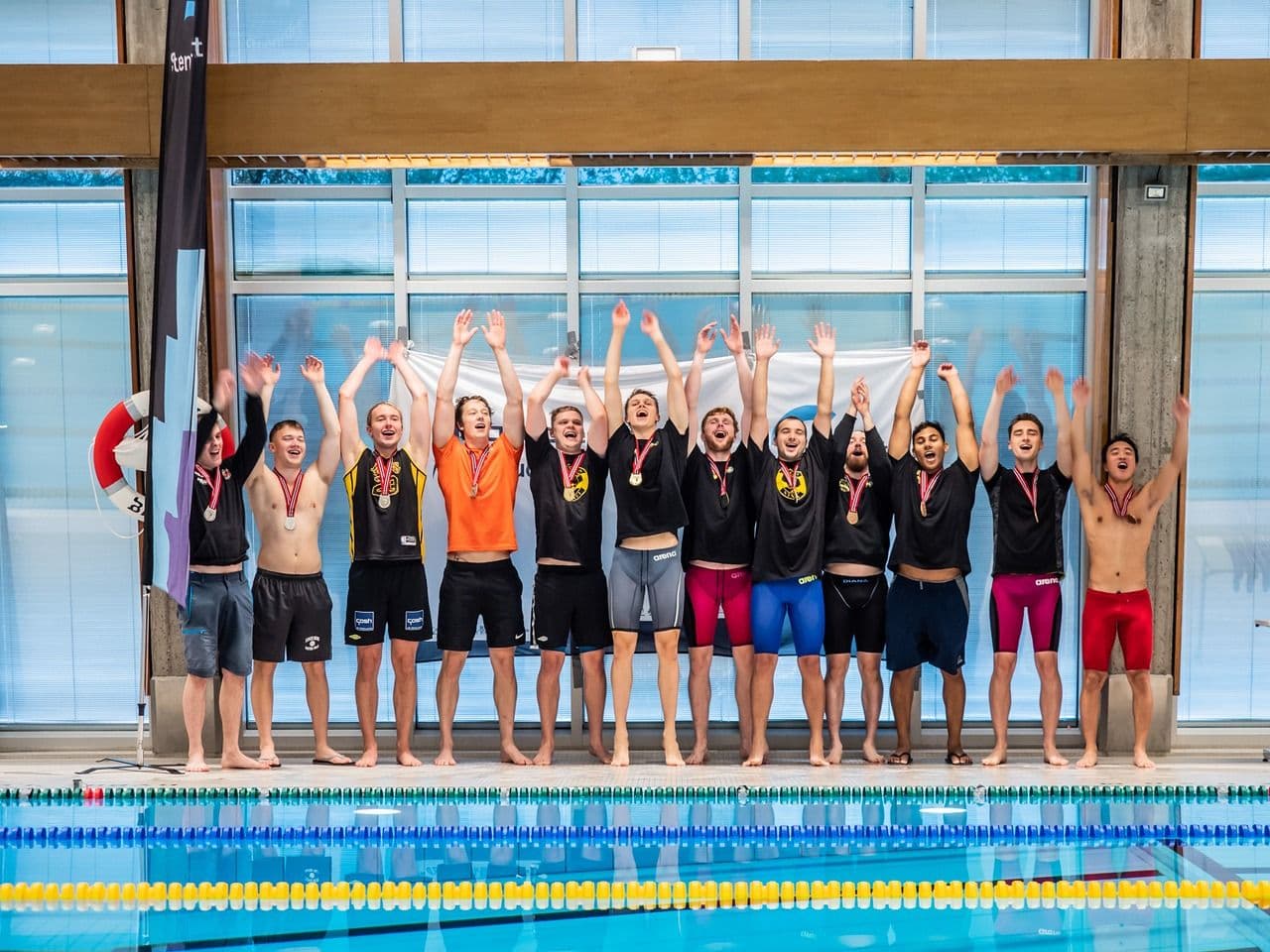 Et lag menn med medaljer jubler med hevede armer ved kanten av et svømmebasseng.