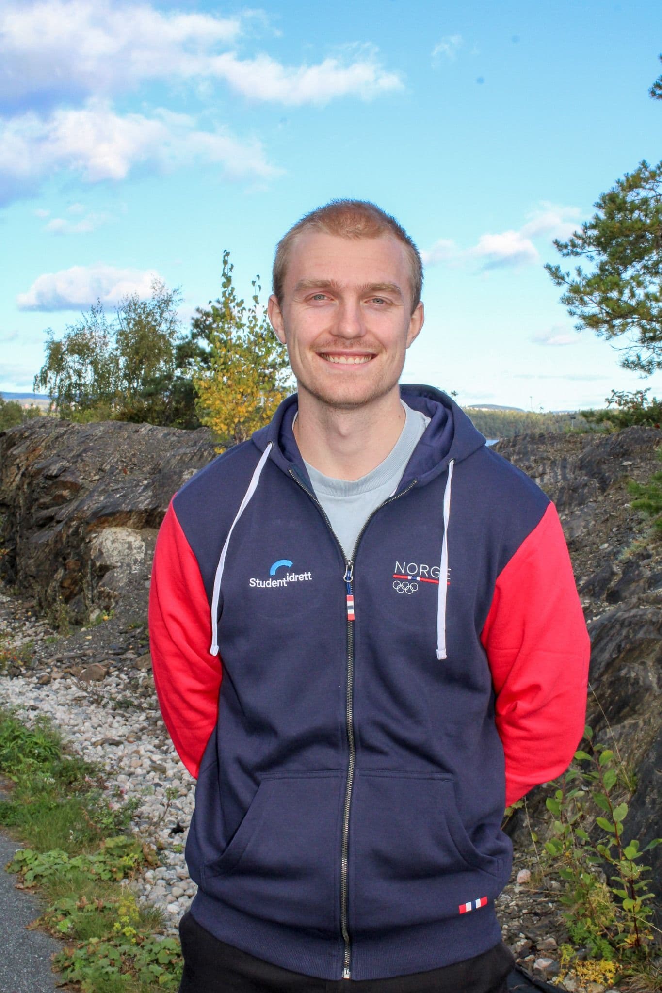 Portrett av en smilende mann i Studentidrett- og Norge-jakke utendørs foran blå himmel.