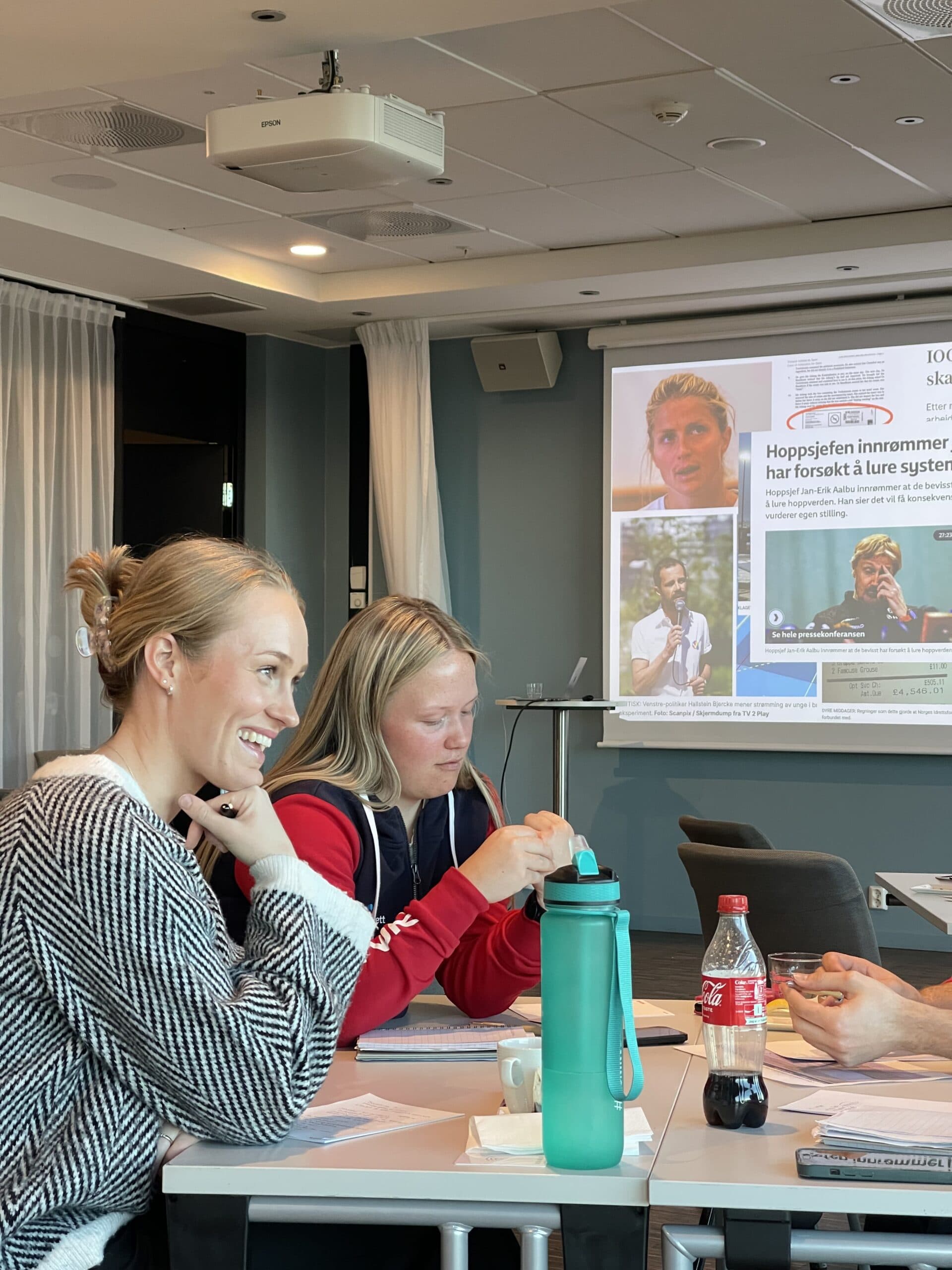 To kvinner sitter og følger med under et seminar der en avisartikkel om hoppsjefen vises på lerretet.