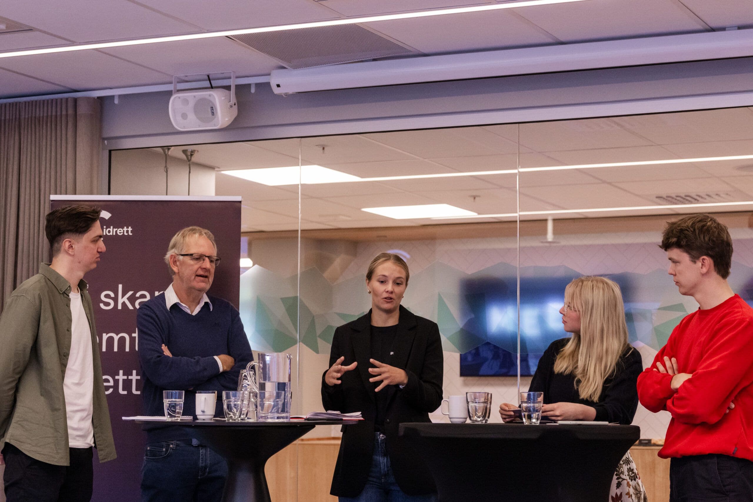 Fem personer deltar i en paneldebatt ved ståbord foran en Studentidrett-rollup.