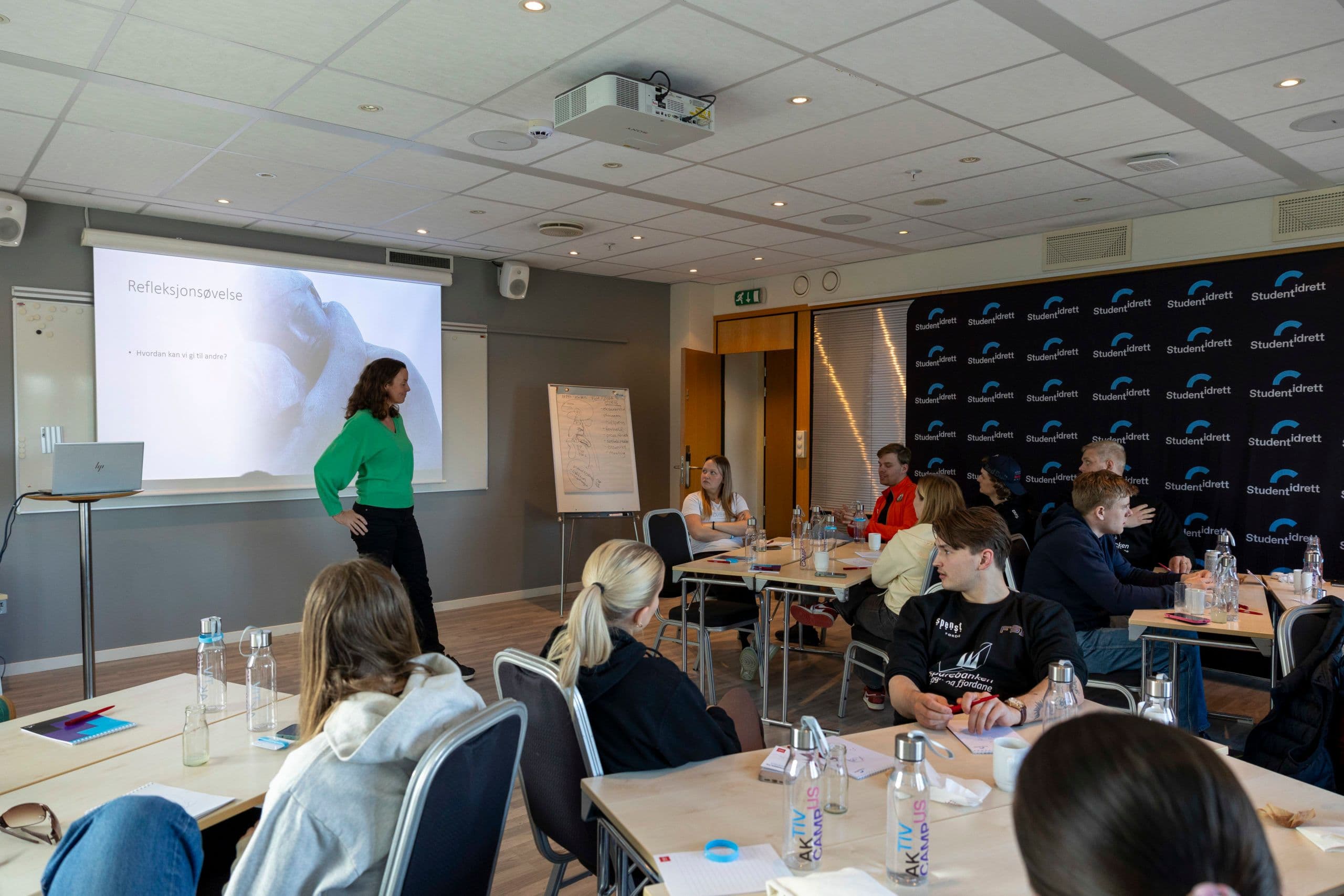 En foredragsholder underviser en gruppe deltakere ved bord i et møterom med Studentidrett-vegg.