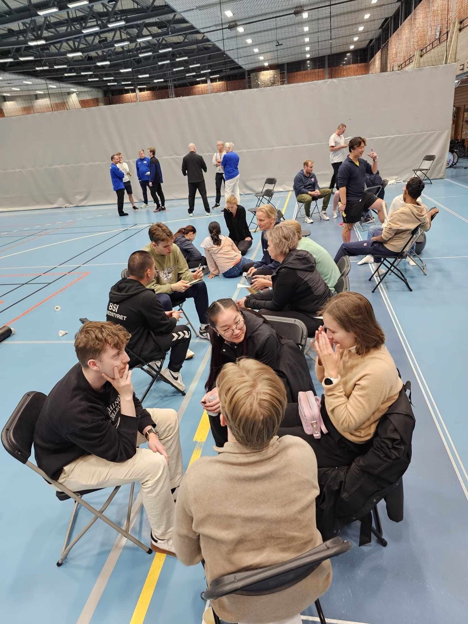 Studenter sitter i mindre grupper på stoler og samtaler i en stor idrettshall.