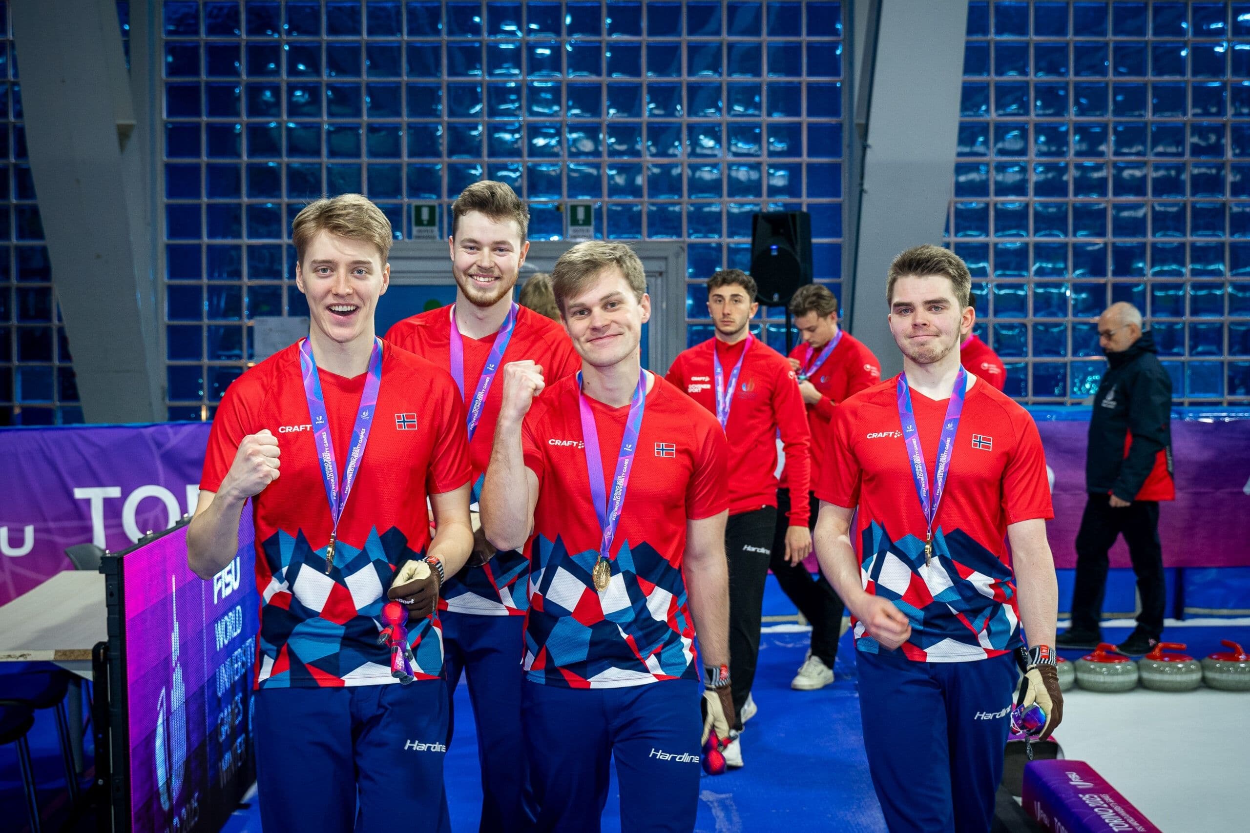 Fire mannlige utøvere i røde drakter feirer med medaljer på en curlingbane foran en FISU-vegg.