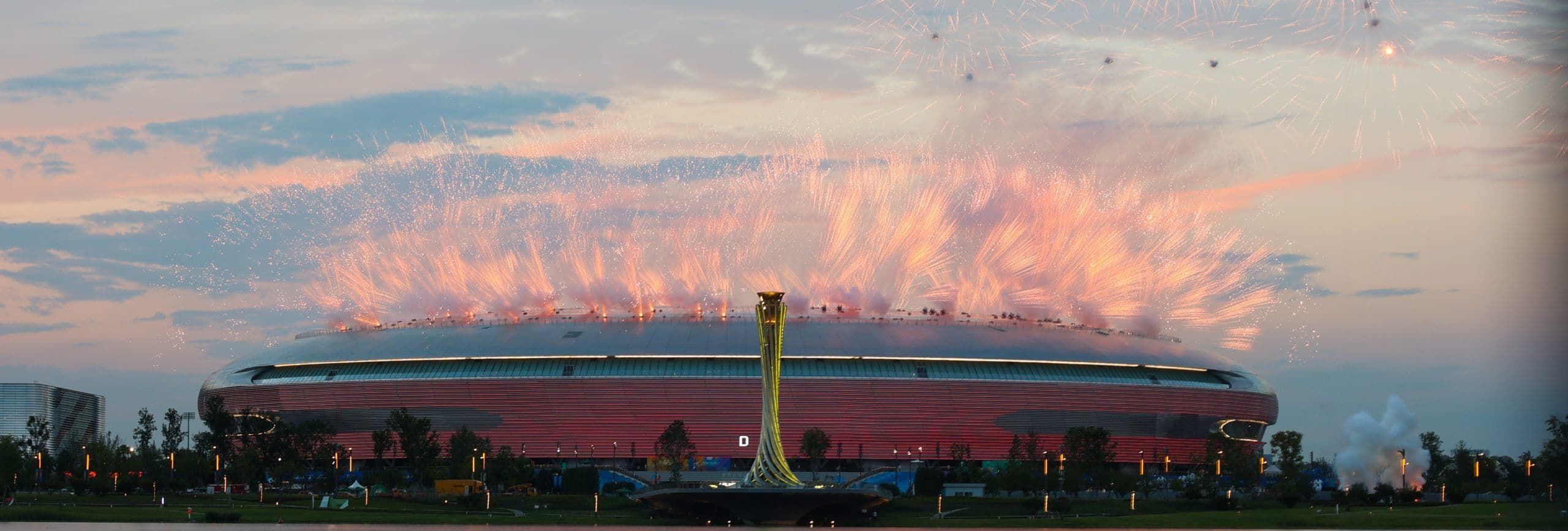 Panorama av et stadion med fyrverkeri over taket under en åpningsseremoni i skumringen.
