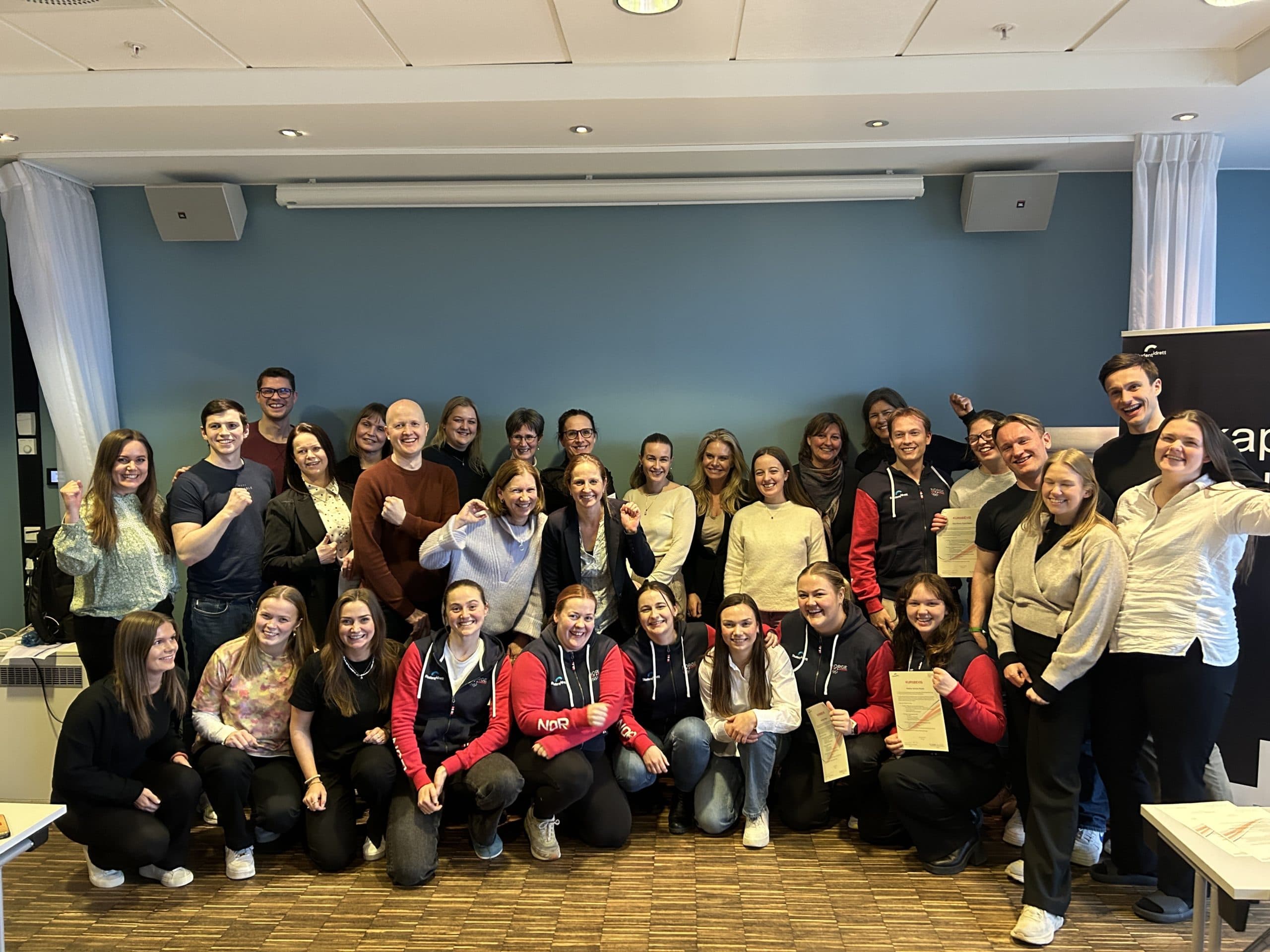 En stor gruppe studenter poserer og jubler samlet i et møterom.