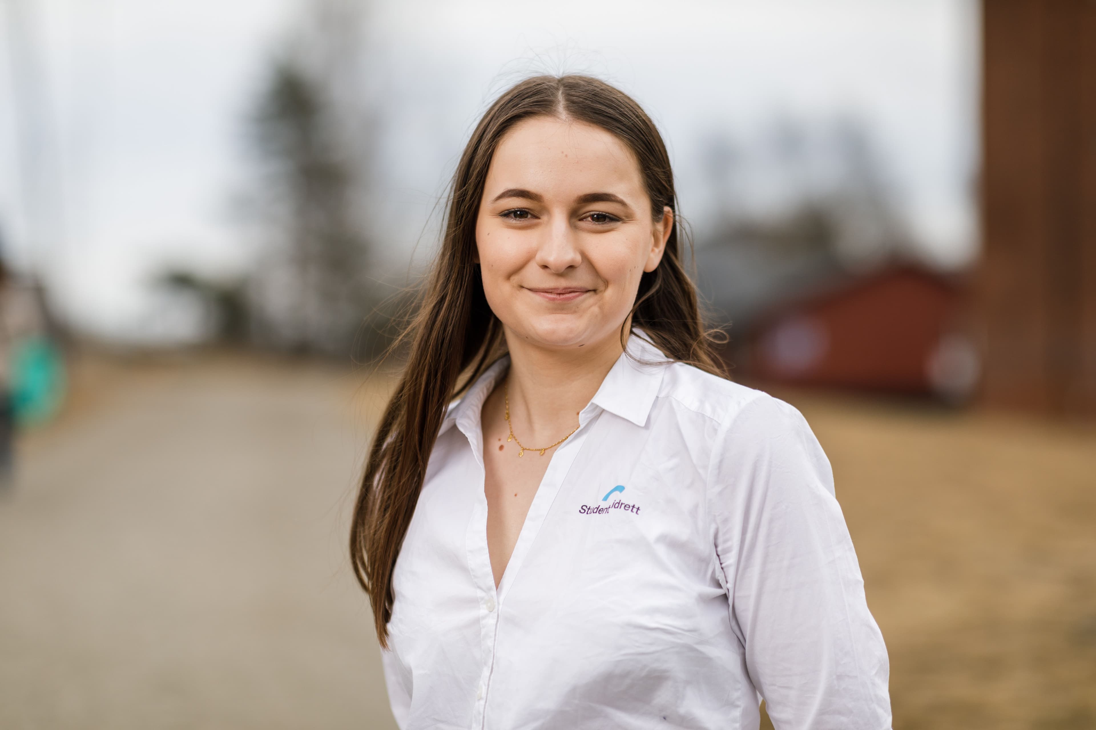 Portrett av en kvinne i hvit skjorte med Studentidrett-logo, fotografert utendørs.