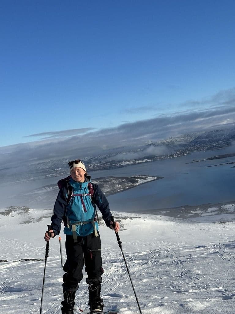 En person på topptur står i snøen med staver og fjord og fjell i bakgrunnen under klar himmel.