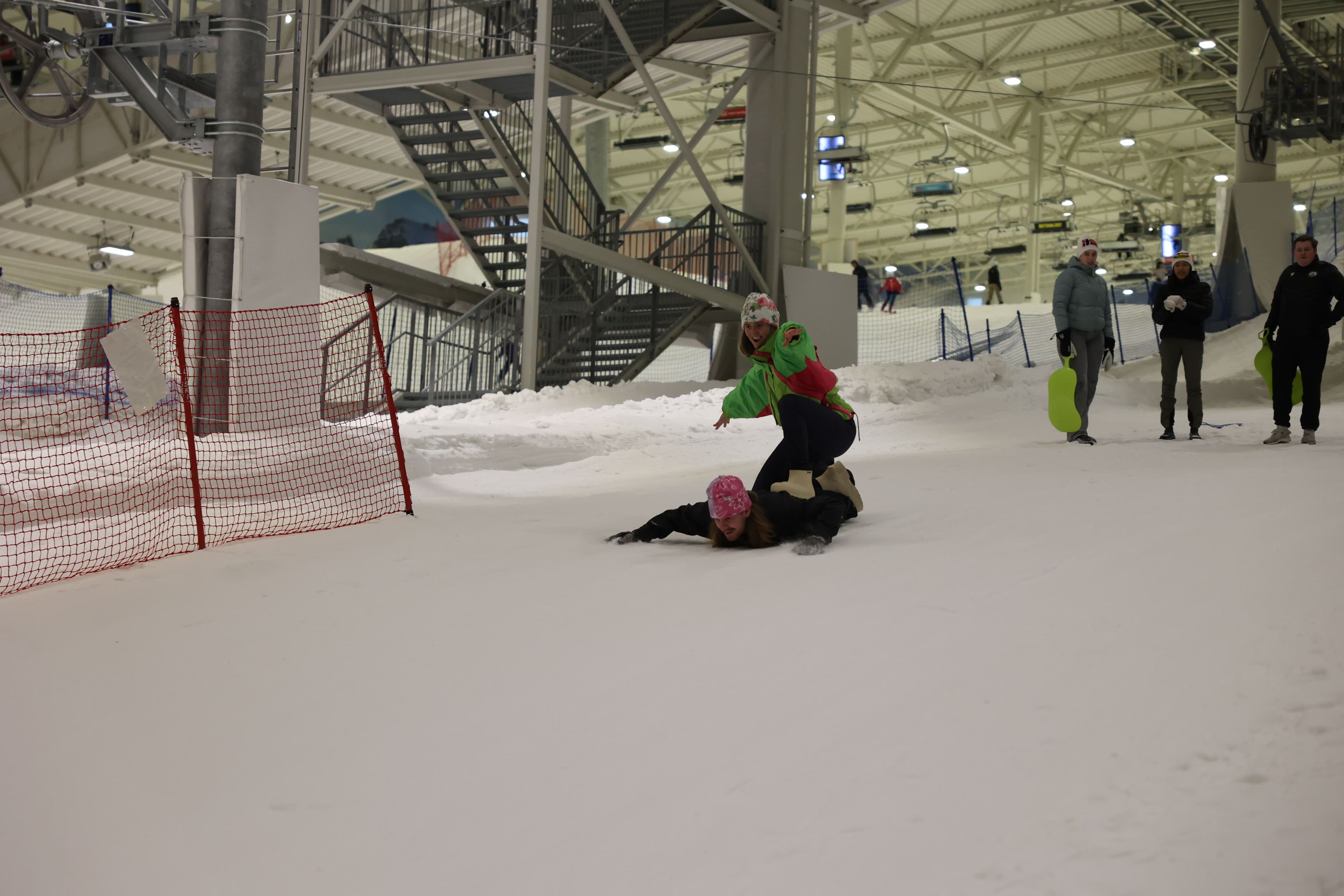 To personer leker og faller i snøen i en innendørs skihall.