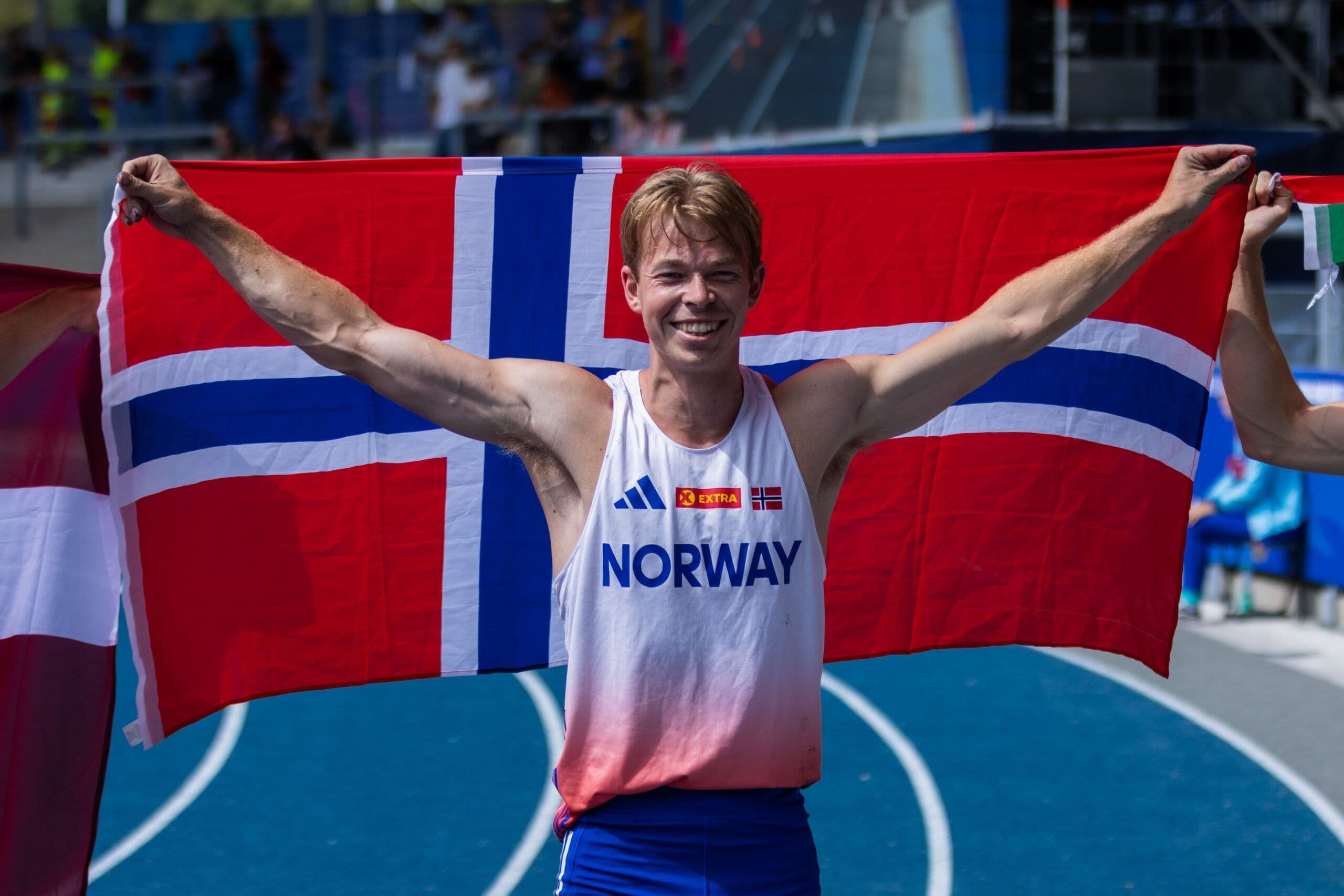 En friidrettsutøver i Norway-drakt smiler og holder opp et norsk flagg på løpebanen.