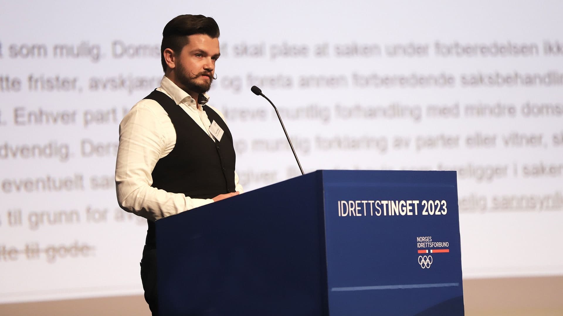 En mann i vest holder innlegg bak en talerstol merket Idrettstinget 2023.