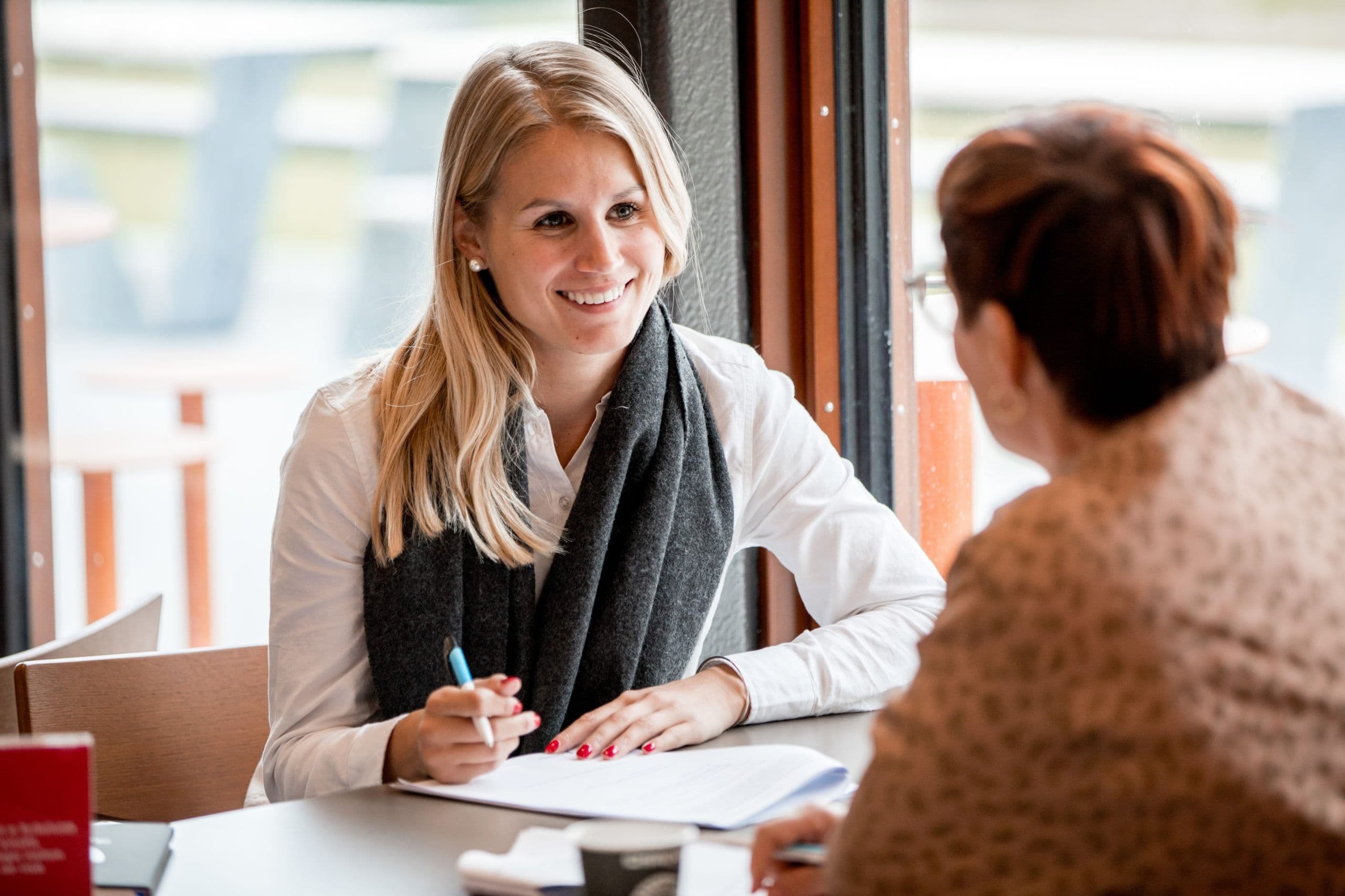 En smilende kvinne sitter ved et bord i en veiledningssamtale med en annen person.