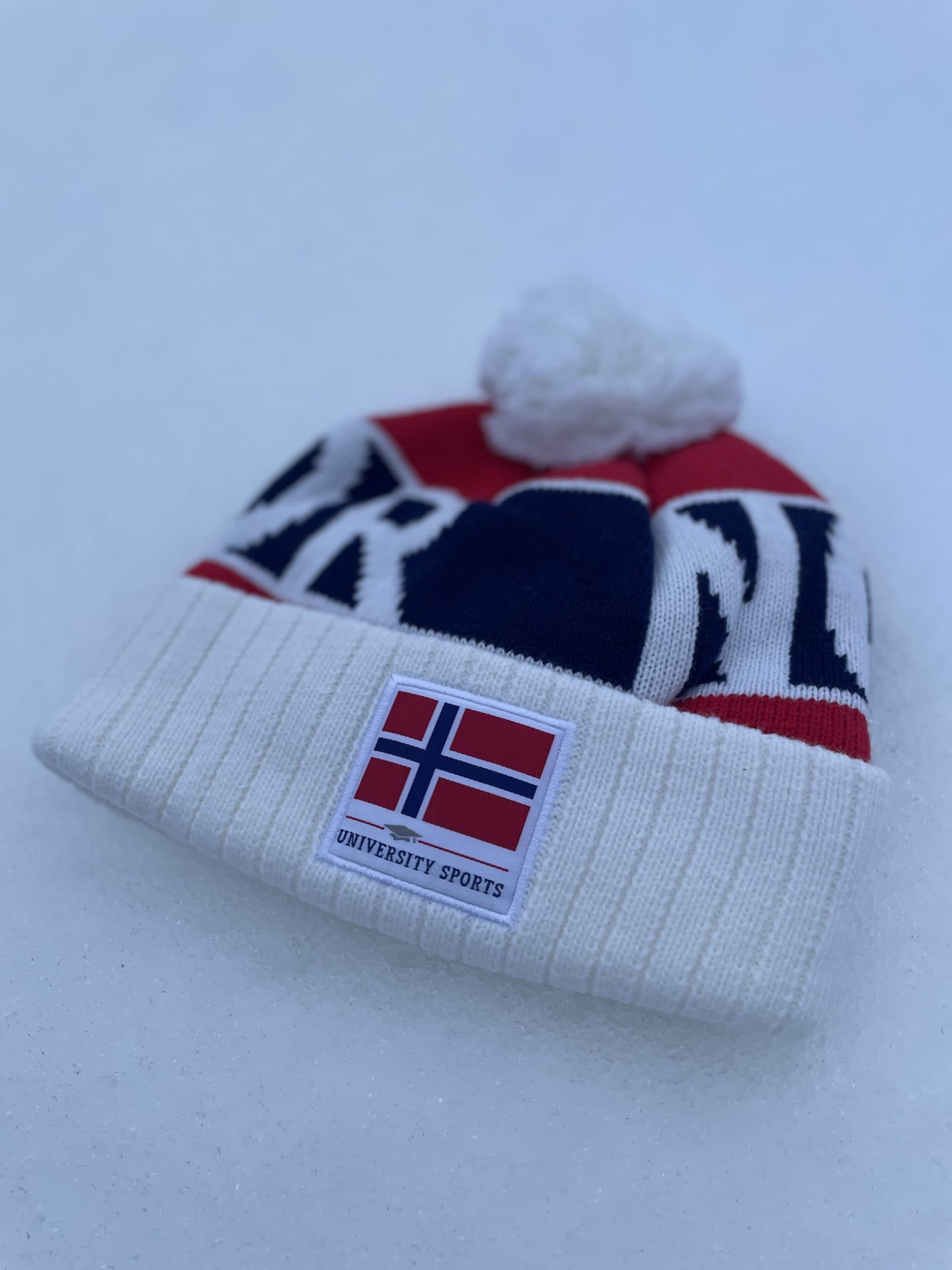 Nærbilde av en hvit pompong-lue med norsk flagg og teksten "University Sports" liggende i snøen.