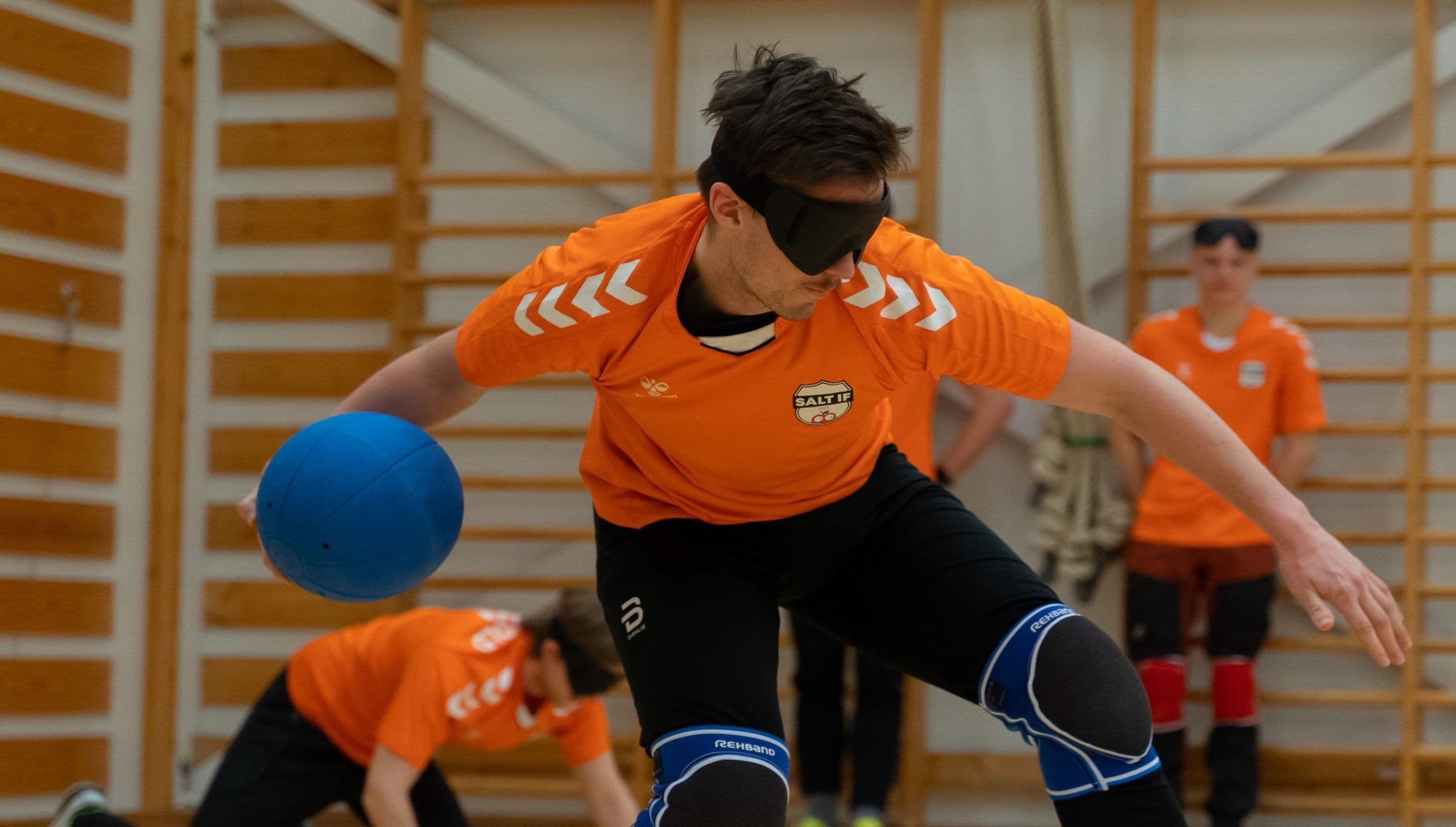 En goalballspiller i oransje drakt med øyemaske forsøker å stoppe en blå ball i en gymsal.