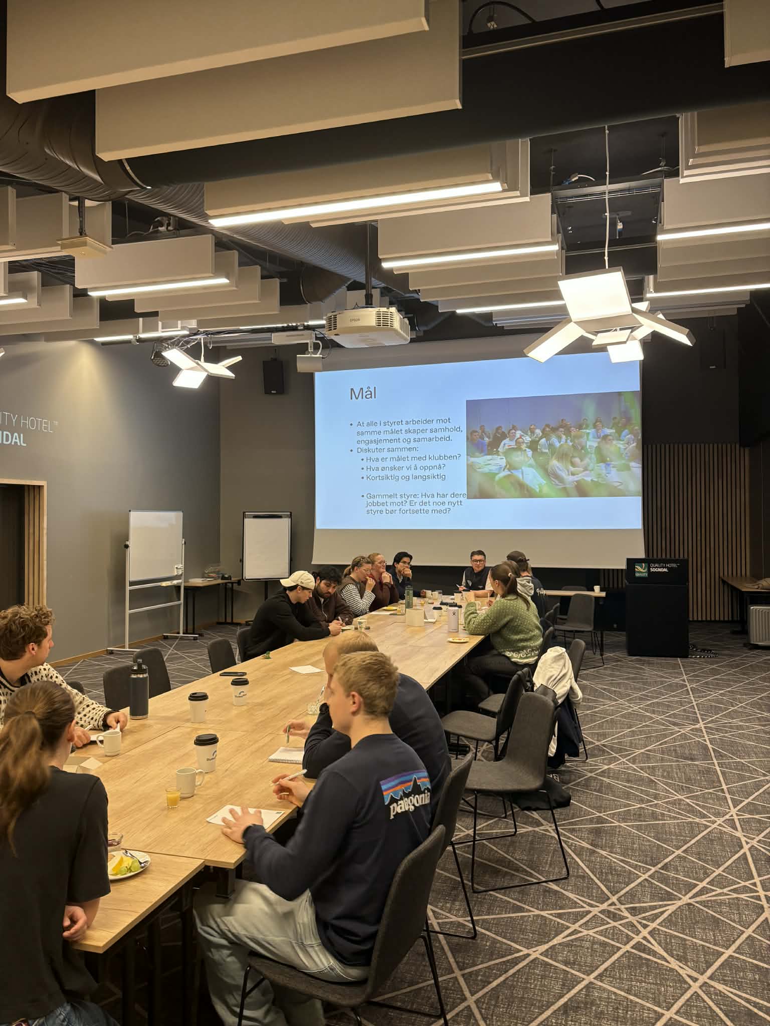 Studenter samlet rundt et langbord i et møterom under et seminar, med en presentasjon med tittelen «Mål» på lerretet.