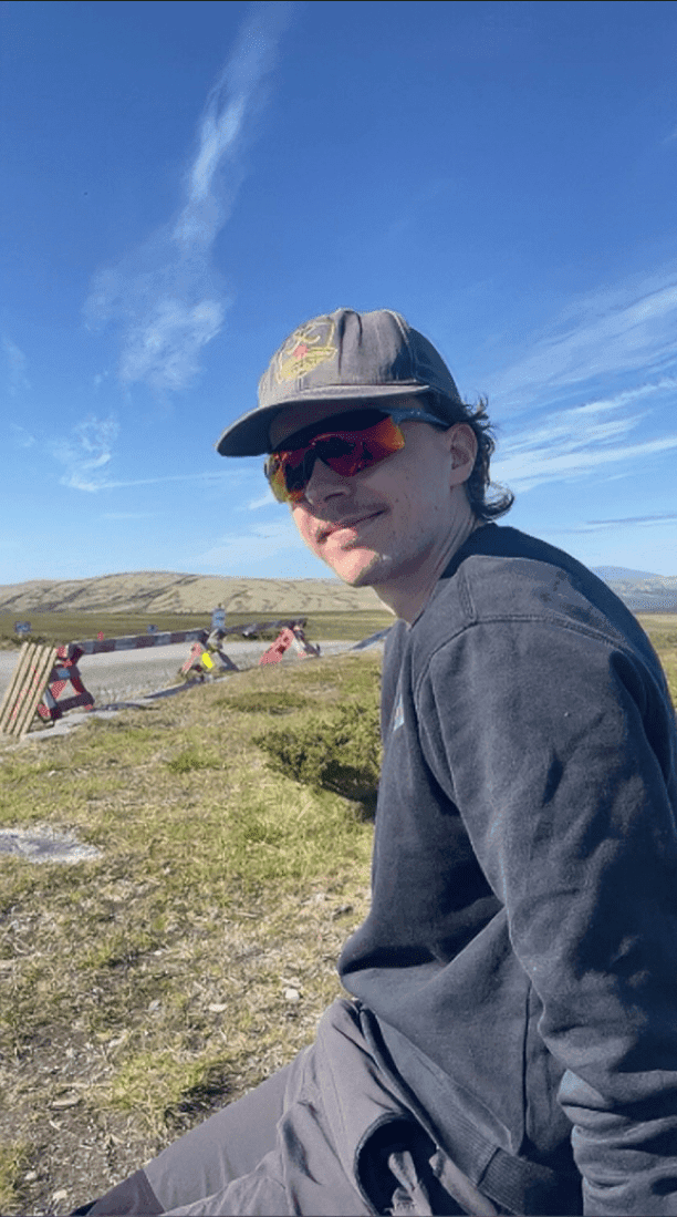 Portrett av en mann med caps og solbriller utendørs i et åpent fjellandskap.