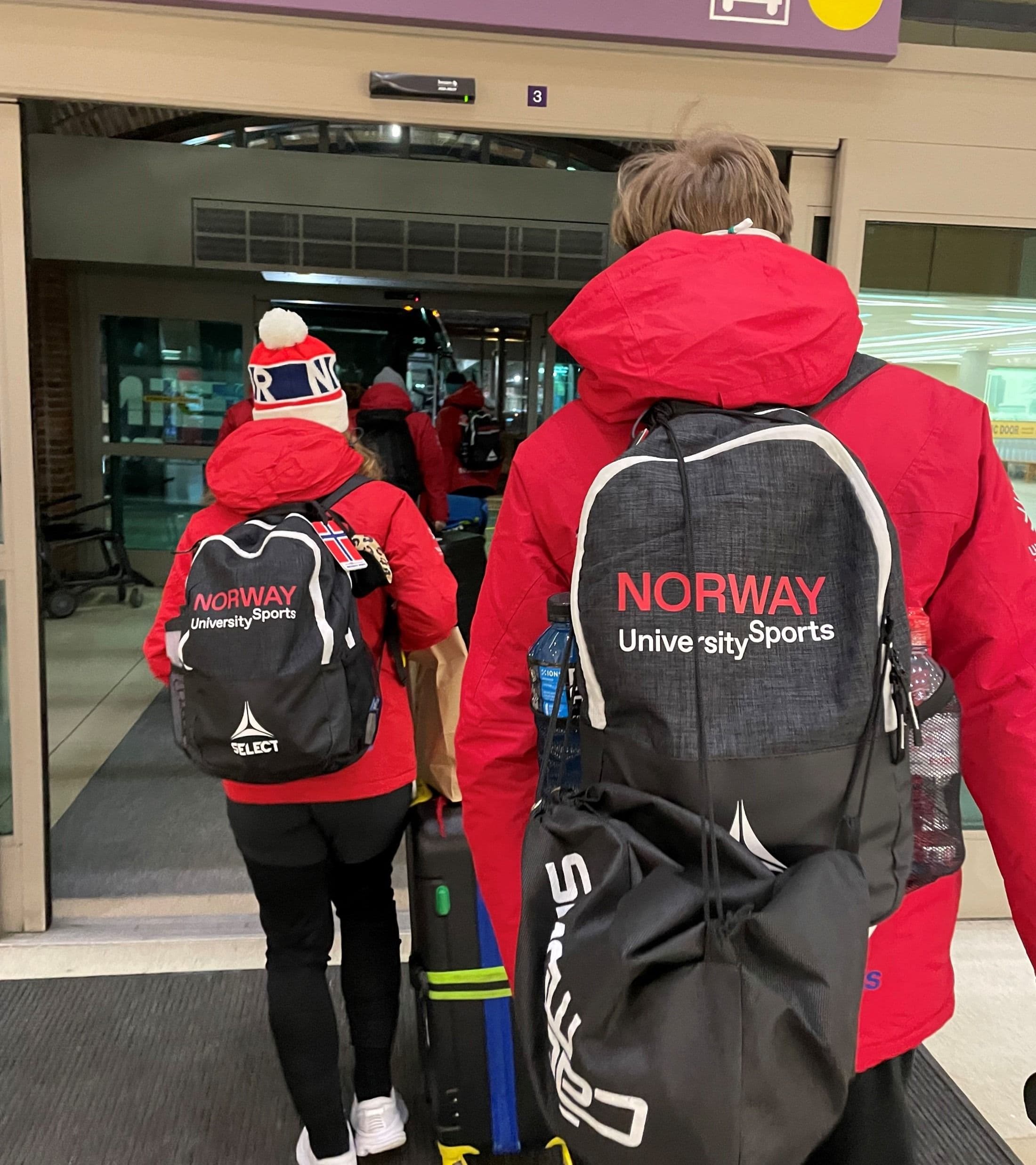 To personer sett bakfra med Norway University Sports-sekker på vei gjennom en flyplassterminal.